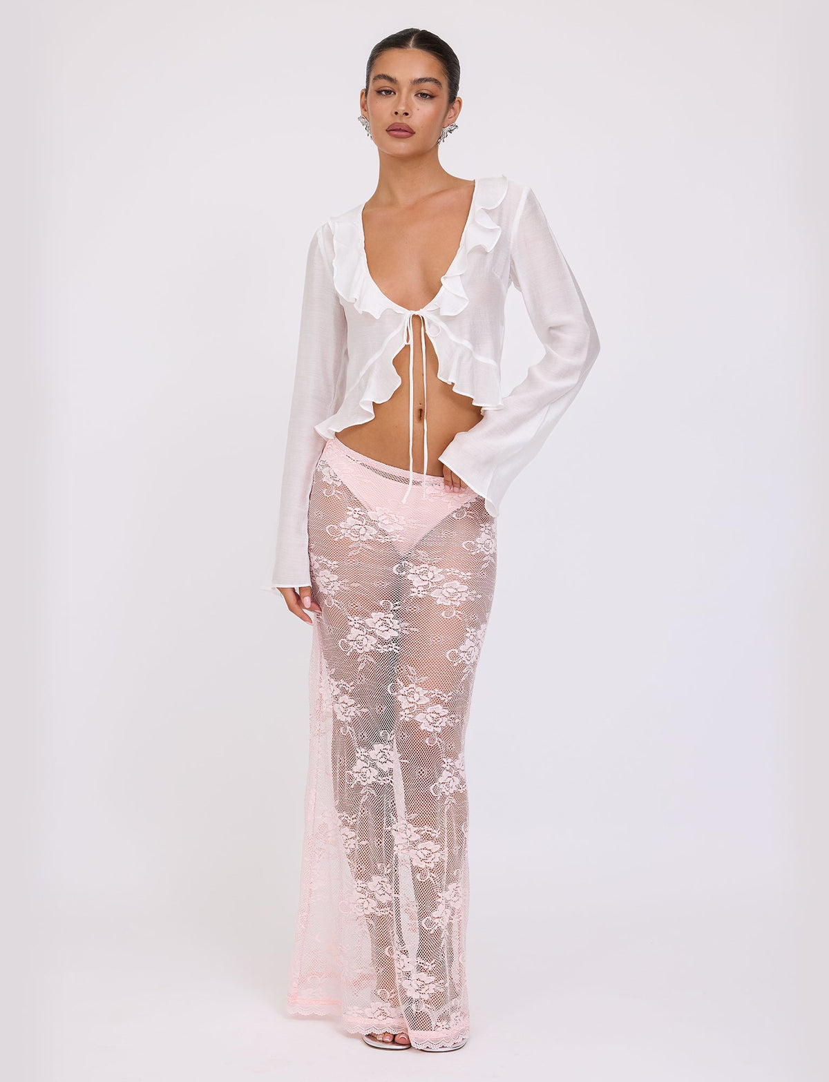 A woman stands against a plain background wearing the CORVIN TOP in white by TIGERMIST. The sheer, long-sleeve top features a deep neckline and ruffled edges. She pairs it with a high-waisted, sheer lace skirt that reveals light-colored shorts underneath. Her hair is pulled back and she complement her outfit with heels.