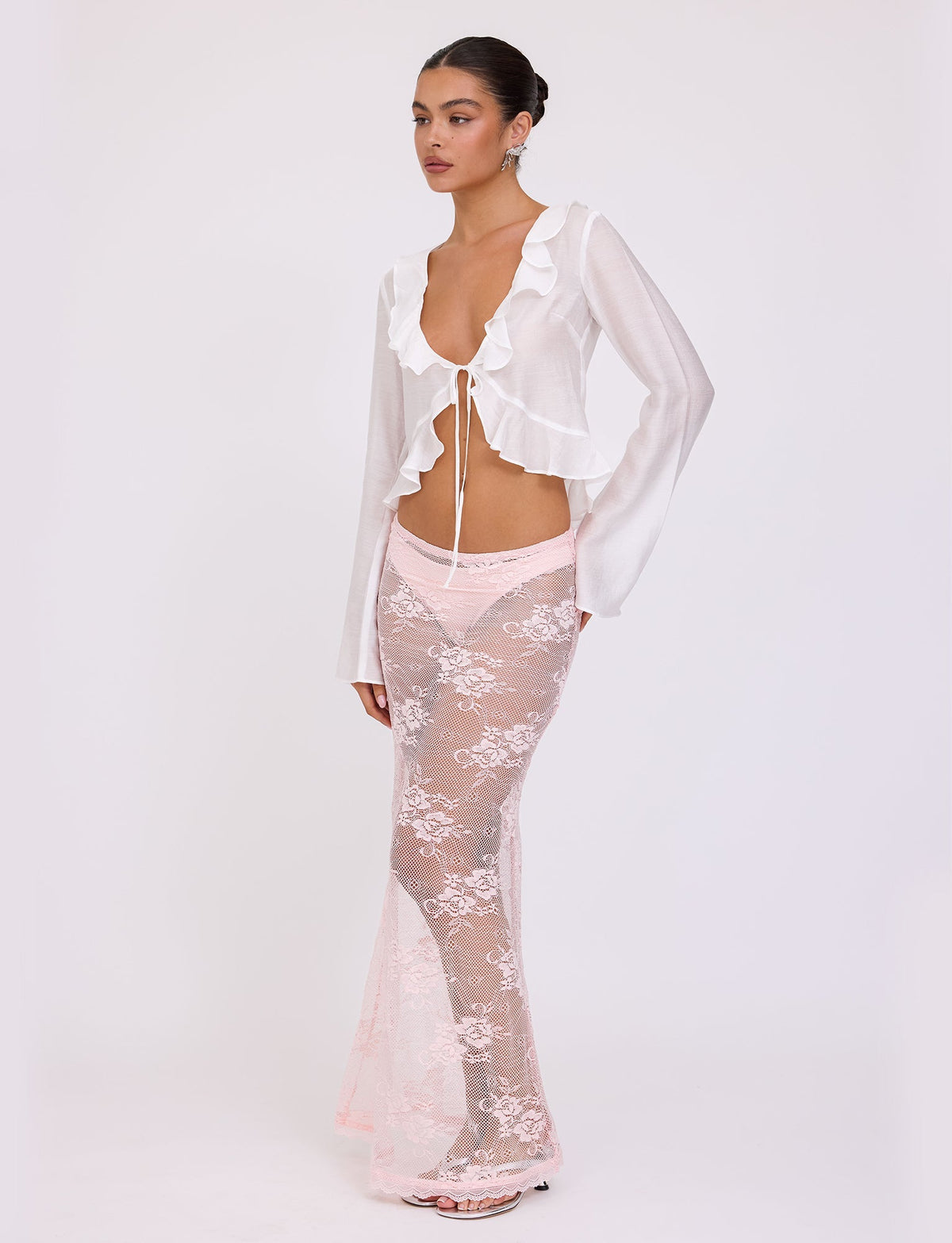 A woman stands against a plain white background, exuding elegance in the TIGERMIST CORVIN TOP - WHITE, a ruffled, long-sleeved top tied at the front. She pairs it with a sheer, lacy, pink maxi skirt worn over pink underwear. Her refined posture and accessories echo the exquisite style of Tiger Mist. Her hair is tied back, completing her chic look.