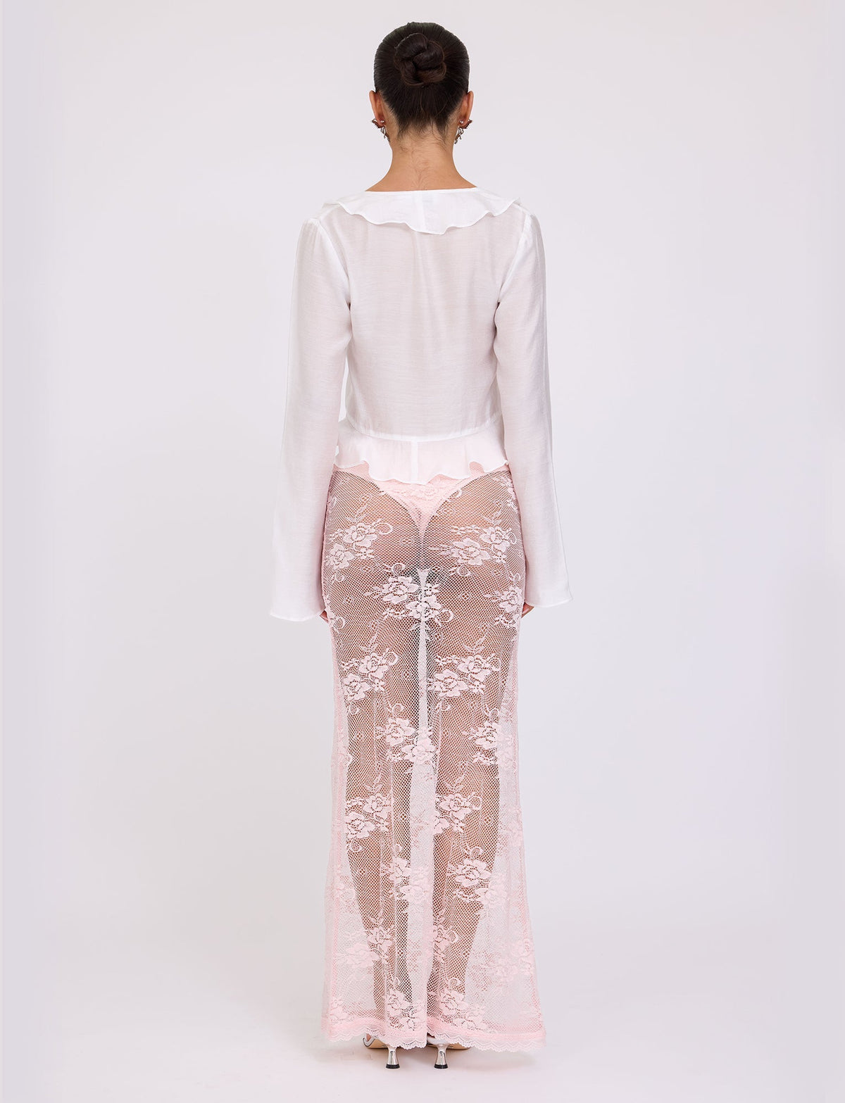 A woman stands with her back to the camera, wearing the CORVIN TOP - WHITE from TIGERMIST paired with pink lace sheer pants. Her hair is tied up in a neat bun, and she completes the look with nude-colored shoes. The image showcases the outfit against a light, plain background.