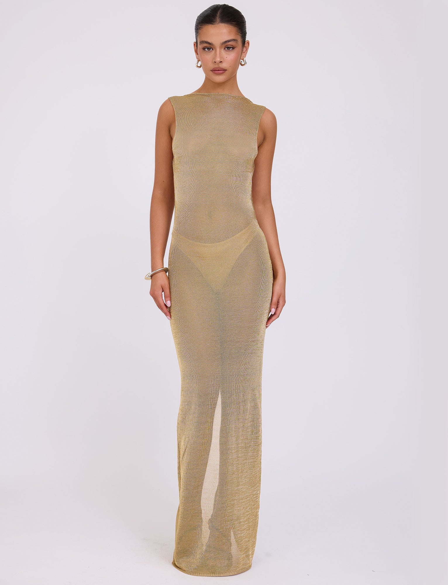 A woman stands against a plain background wearing the sleeveless, long, semi-sheer ZAELA DRESS in gold from TIGERMIST. She has her hair pulled back and accessorizes with earrings and a bracelet. The dress features a high neckline and is fitted to her body.