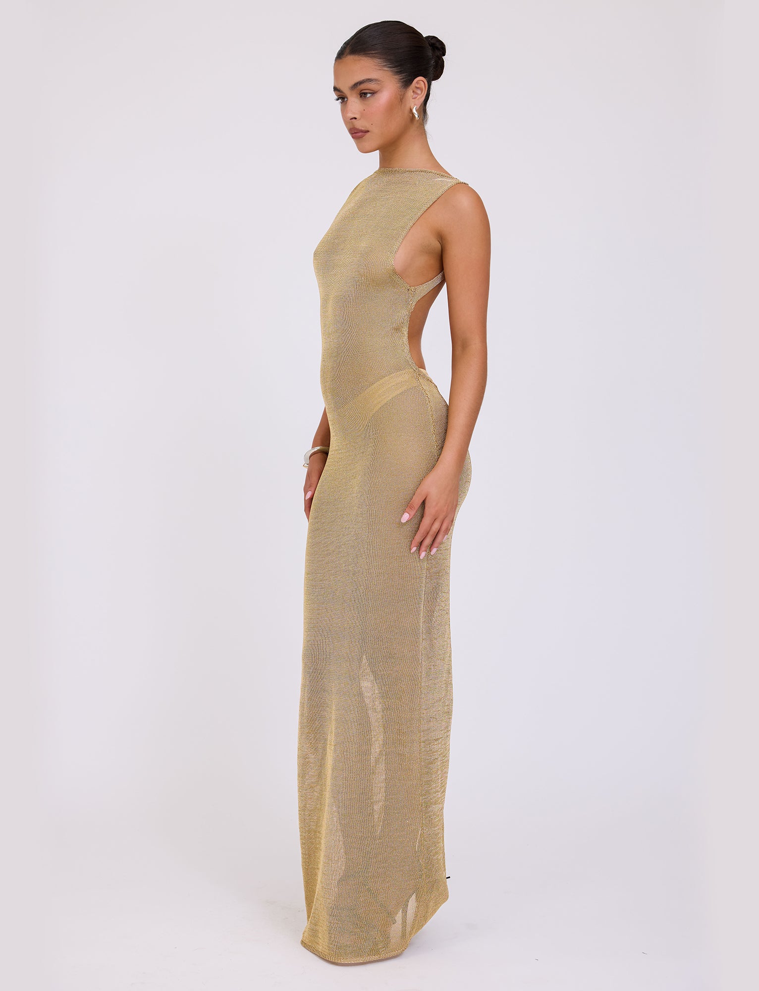 A woman with her hair in a bun poses sideways against a plain white background, wearing the TIGERMIST ZAELA DRESS - GOLD. The dress is long and shimmery, fitted and sleeveless with a high neckline. She accentuates the look by lightly touching her hand to the side of her thigh.