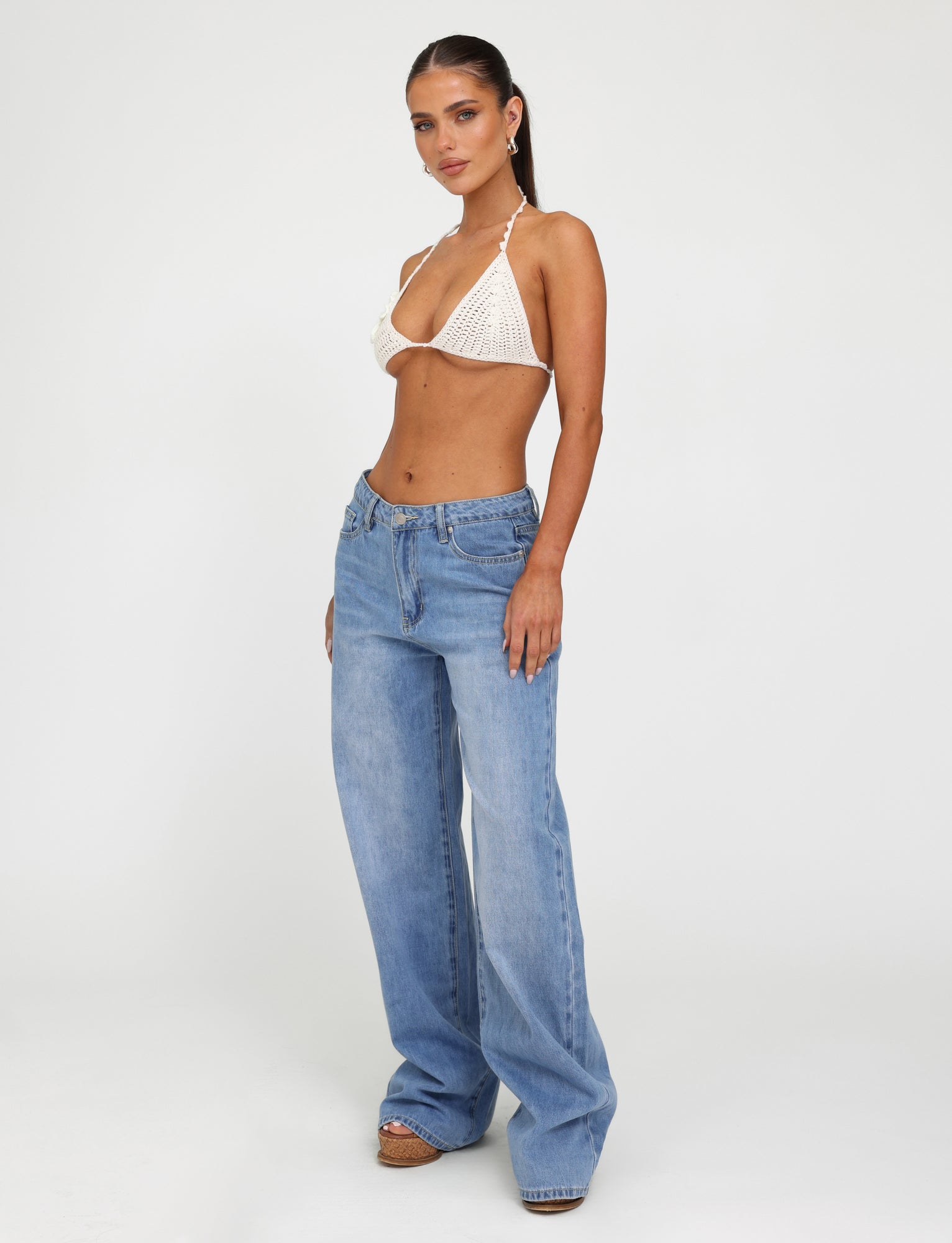 A woman stands confidently against a plain white background, dressed in a white crocheted bikini top from Tiger Mist and the AVEREE JEAN - BLUE : MID WASH relaxed-fit jeans by TIGERMIST, which sit low on her hips. Her hair is pulled back, and she gazes towards the camera with a relaxed expression.