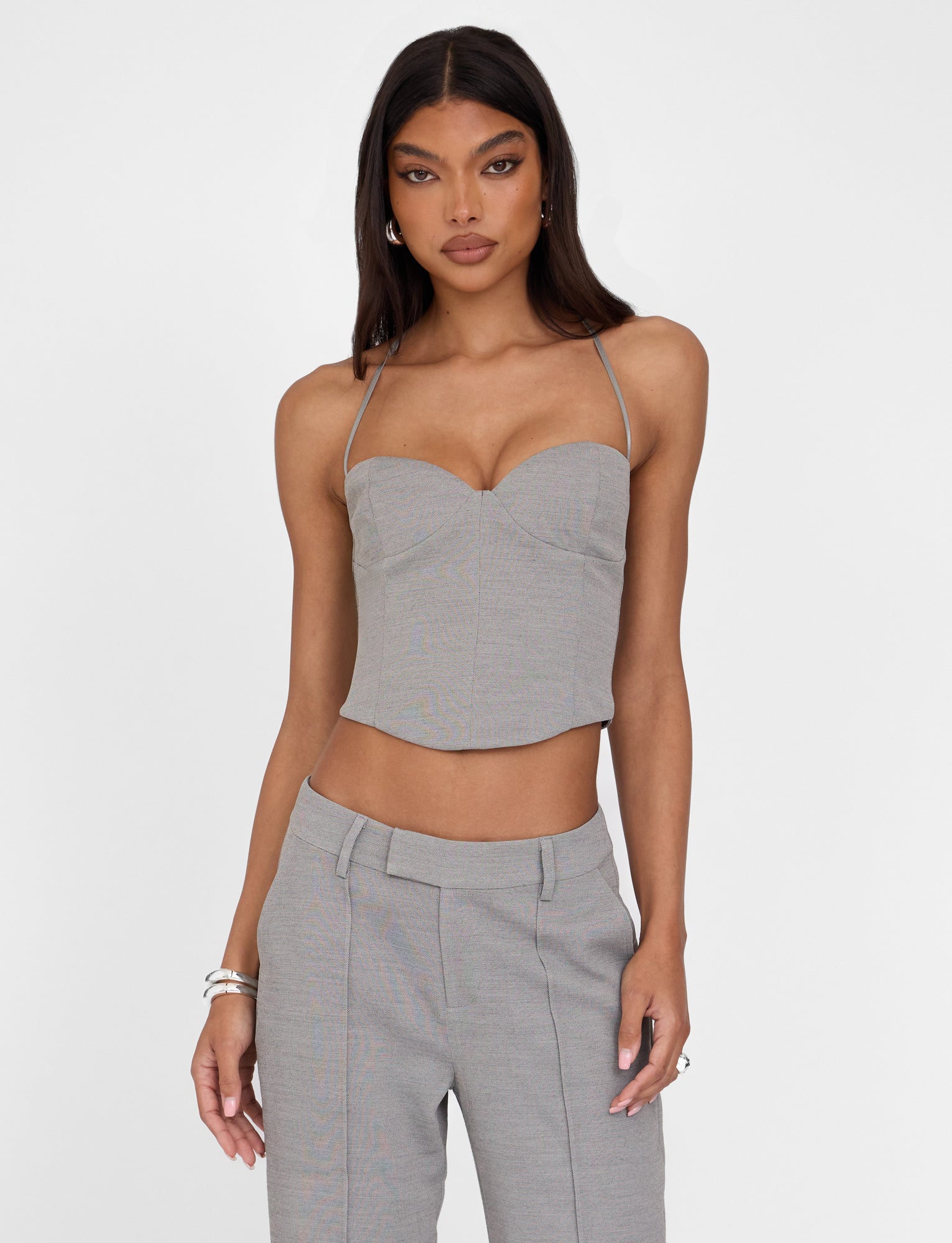 A woman with long dark hair is wearing a fitted TIGERMIST MCKAY TOP - GREY with thin straps and matching high-waisted pants. She has her left hand in her pocket and is accessorized with silver bracelets on her right wrist and hoop earrings, set against a plain white background.