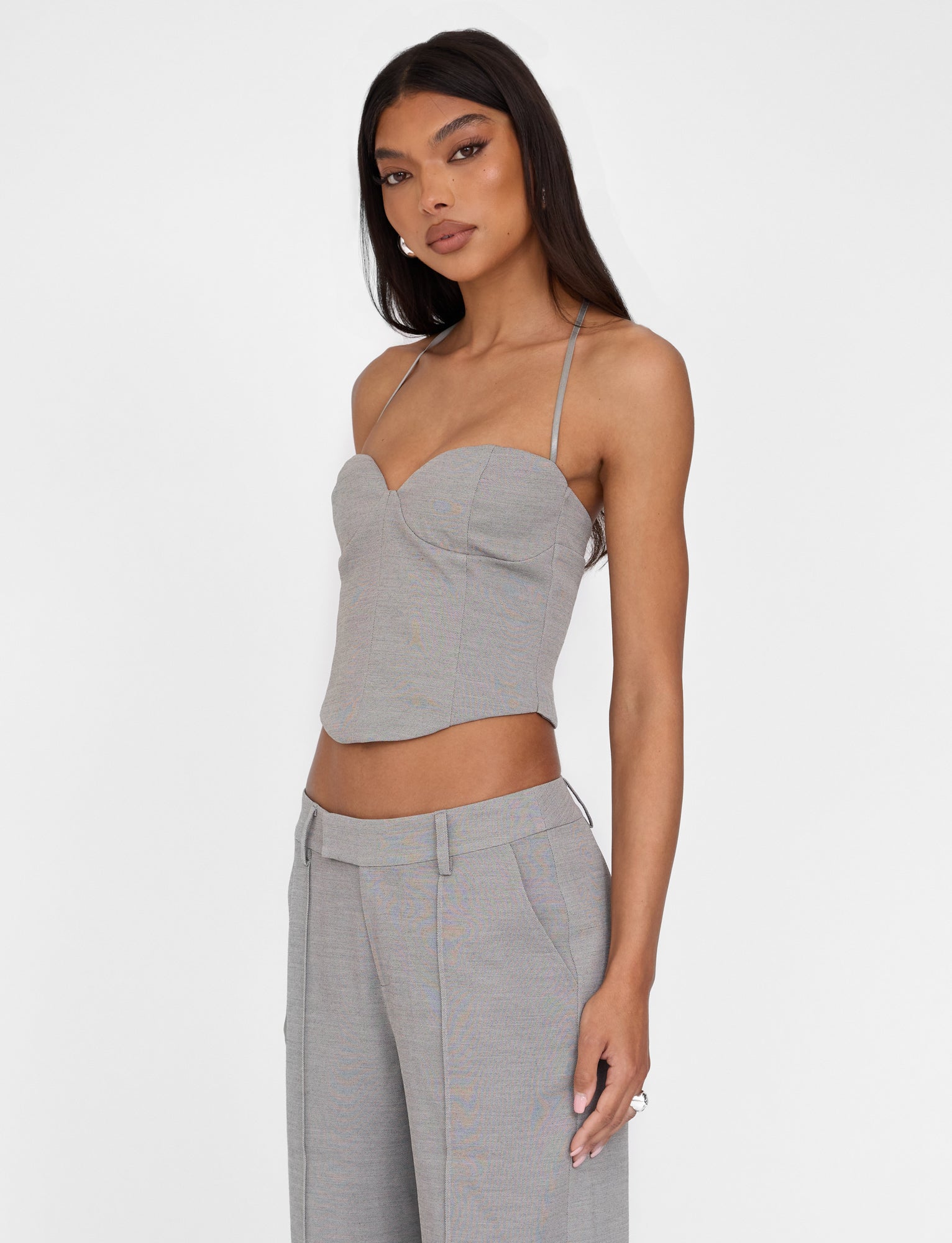 A woman with long, straight black hair is wearing the TIGERMIST MCKAY TOP in grey, featuring an adjustable halter neck strap and paired with matching grey wide-leg trousers. She stands confidently against a plain white background, looking slightly to her left. Her outfit has a tailored, fitted appearance.