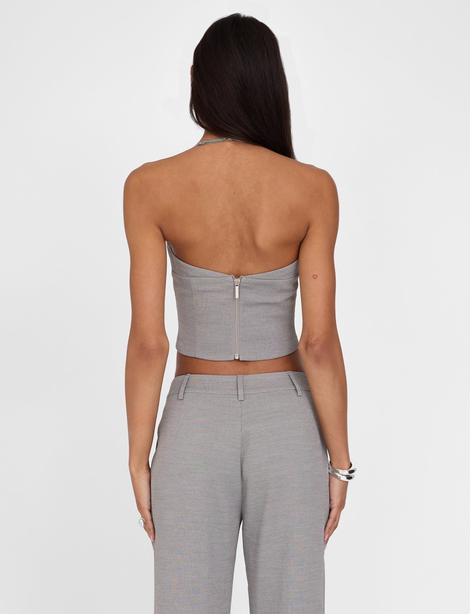 A woman with long dark hair is seen from the back, wearing the TIGERMIST MCKAY TOP in grey, which features a strapless design and a tailored bodice. She pairs it with matching high-waisted pants. Several silver bracelets adorn her left wrist. The background is plain white.