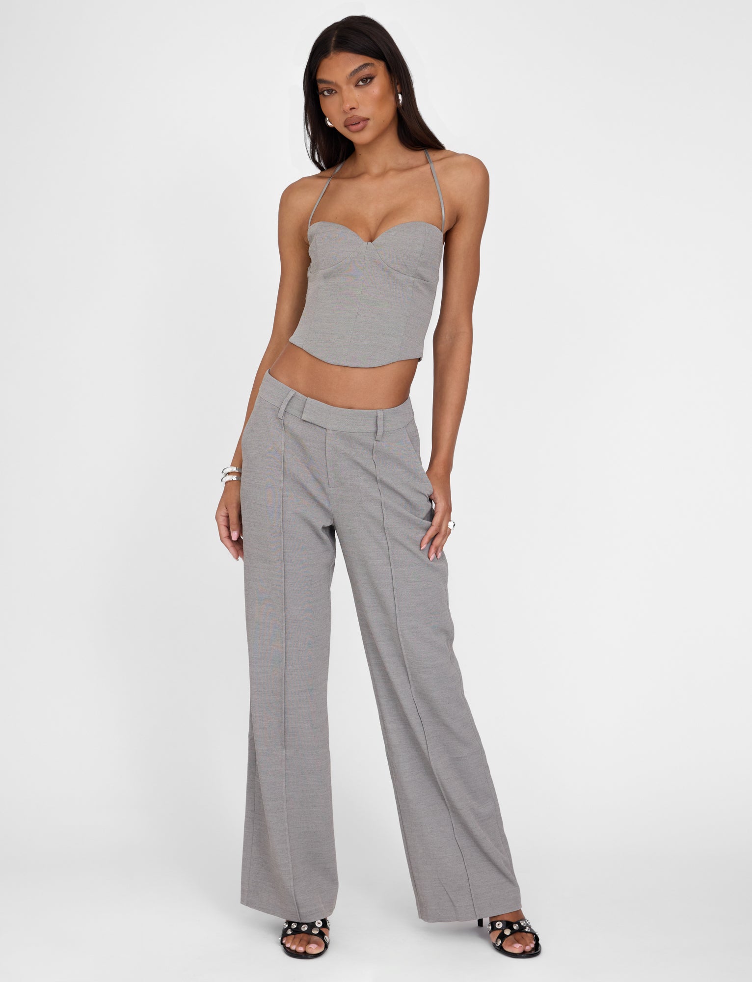 A woman stands against a plain white background, wearing the TIGERMIST MCKAY TOP in gray with a tailored bodice and matching wide-leg pants. She has long, straight hair and accessorizes with bracelets on one wrist and black open-toe heels.
