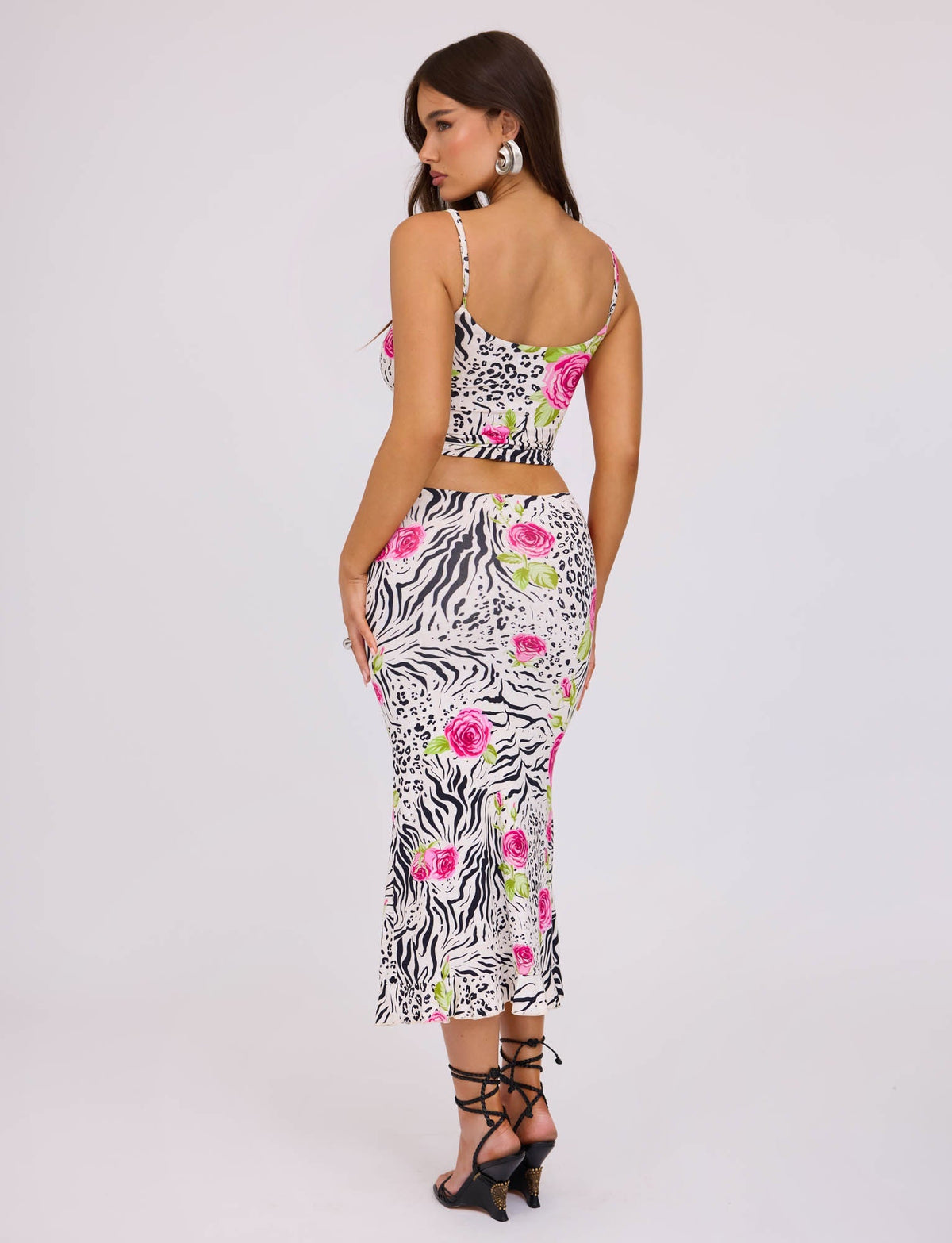 A woman with long brown hair stands facing sideways, wearing a stylish ensemble from TIGERMIST. Her outfit includes a spaghetti strap crop top paired with the IRIE SKIRT - MULTI : ZEBRA, which showcases a zebra print pattern adorned with pink and green floral accents. She completes her look with black strappy heels.