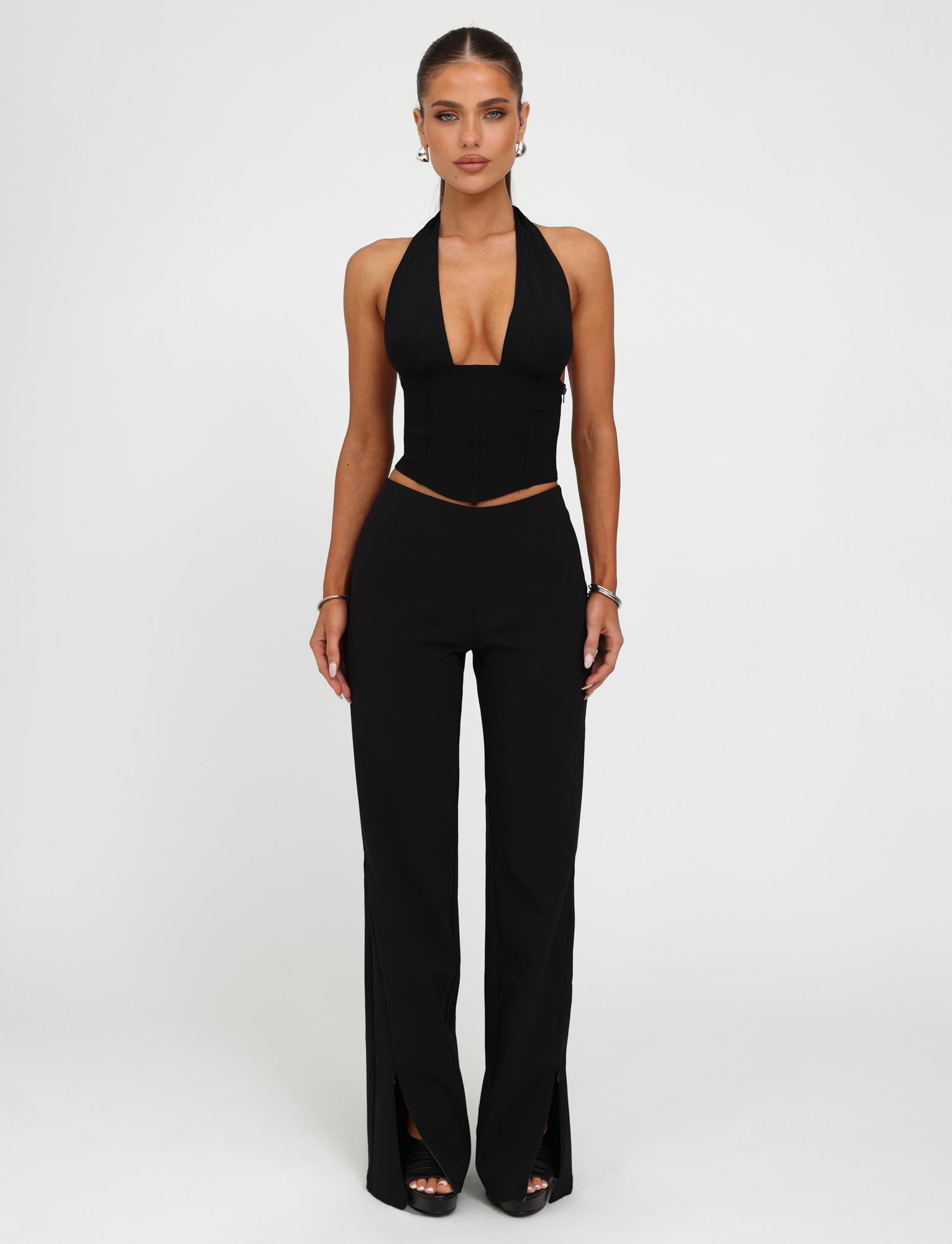 A woman stands against a white background wearing the TIGERMIST JANECE TOP in black, a corset-style halter-neck with a deep V neckline, paired with high-waisted black pants with front slits. Her hair is tied back and she accessorizes with earrings and bracelets.