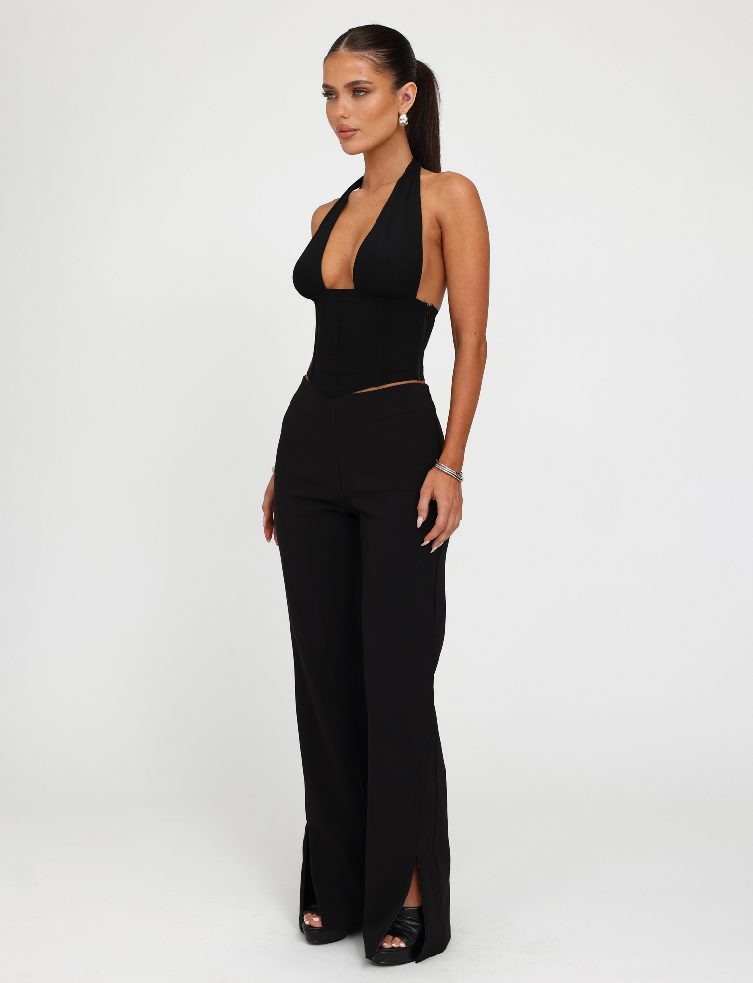 A woman in a ponytail poses against a white background wearing the TIGERMIST JANECE TOP - BLACK, a corset style deep V-neck, paired with high-waisted wide-leg black pants with front slits and open-toe heels.