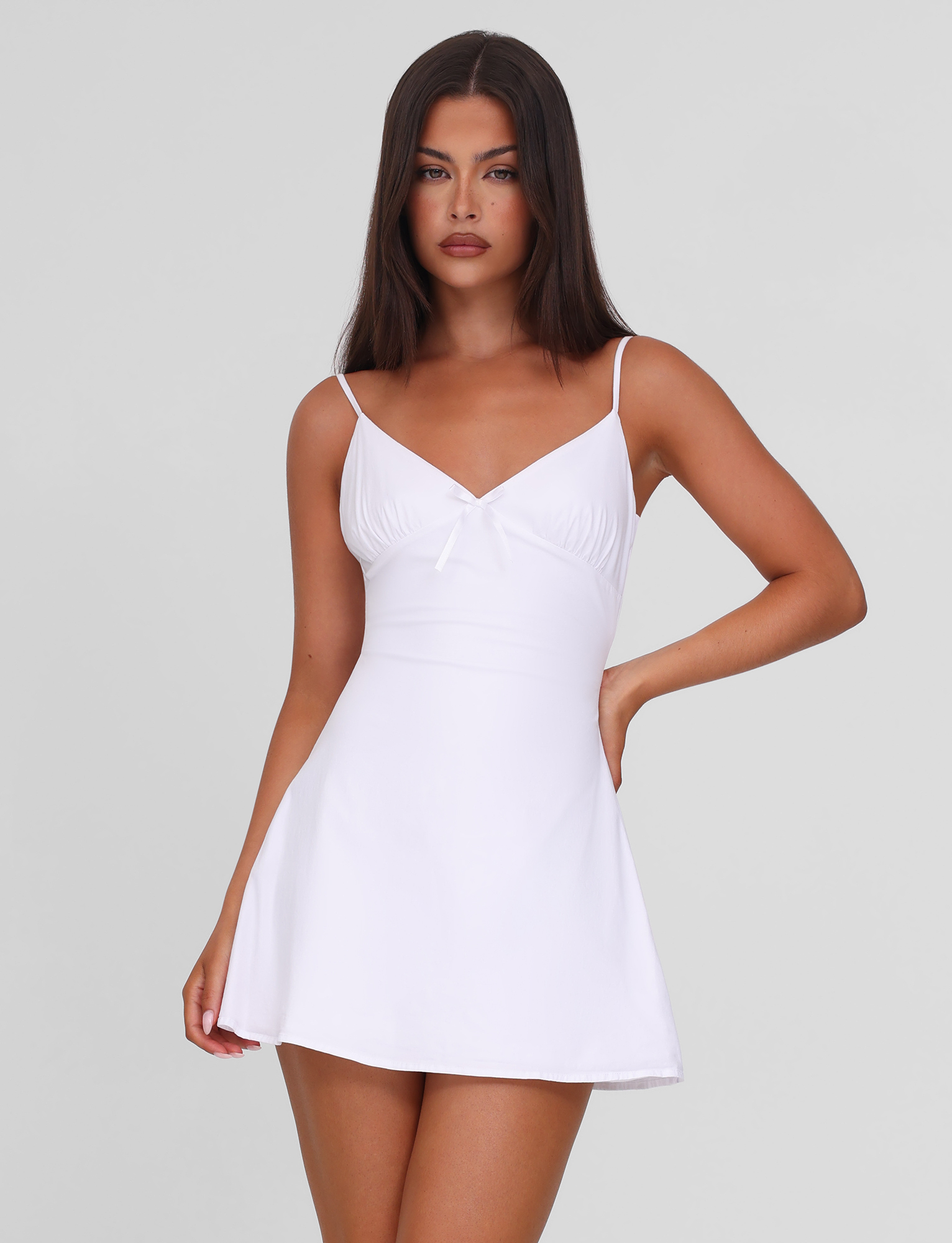 A woman with long dark hair models the TIGERMIST WINNIE MINI DRESS in white, featuring adjustable shoulder straps. She stands against a plain light gray background, one hand on her hip and the other by her side.
