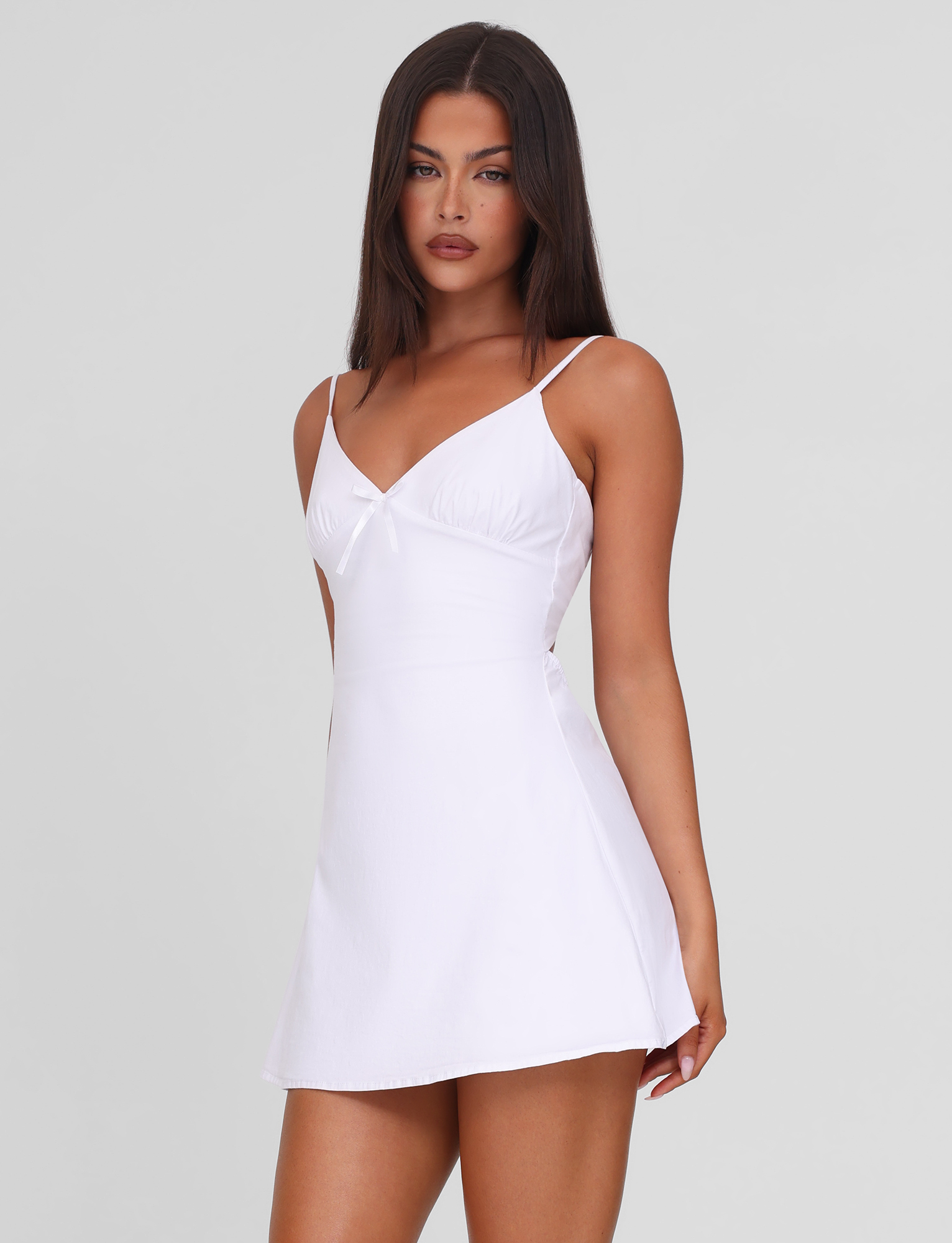 A woman with long, straight dark hair wears the TIGERMIST WINNIE MINI DRESS in white, featuring a V-neckline, central bow detail, and adjustable straps. She stands against a light gray background, gazing slightly to the side.