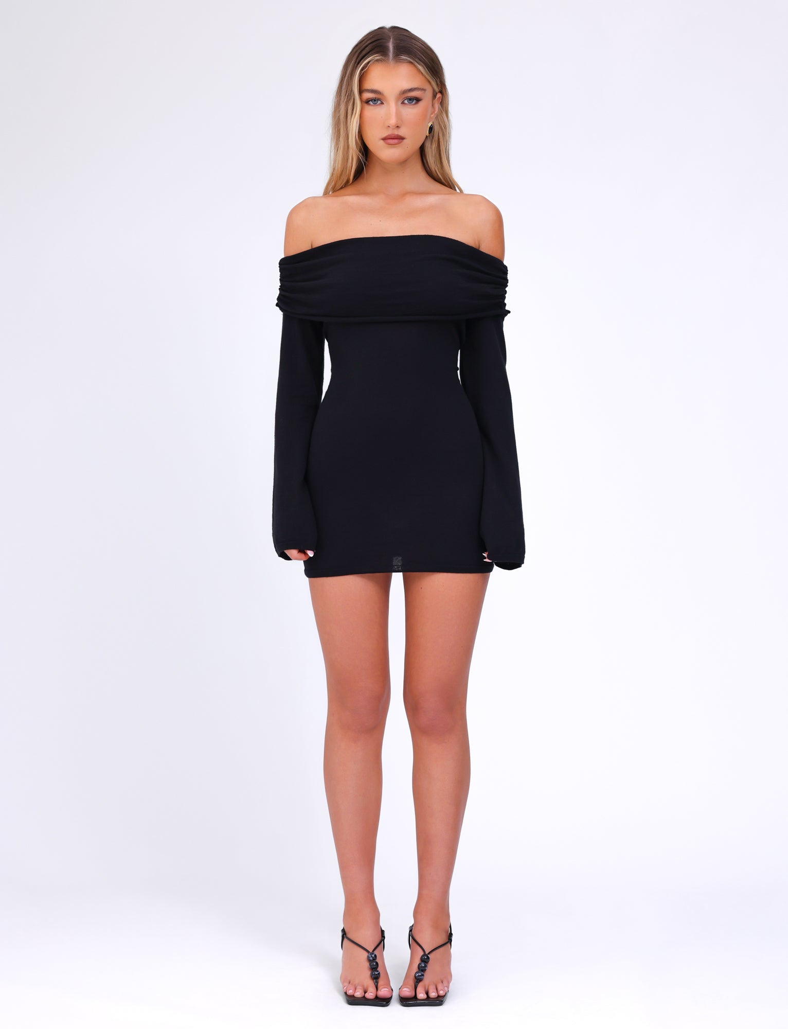 A woman stands against a plain white background, wearing the off-the-shoulder, long-sleeved DIEDRE JUMPER DRESS - BLACK by TIGERMIST, which features a fitted silhouette. She completes her outfit with black strappy heeled sandals. She has long, straight hair and a neutral expression.