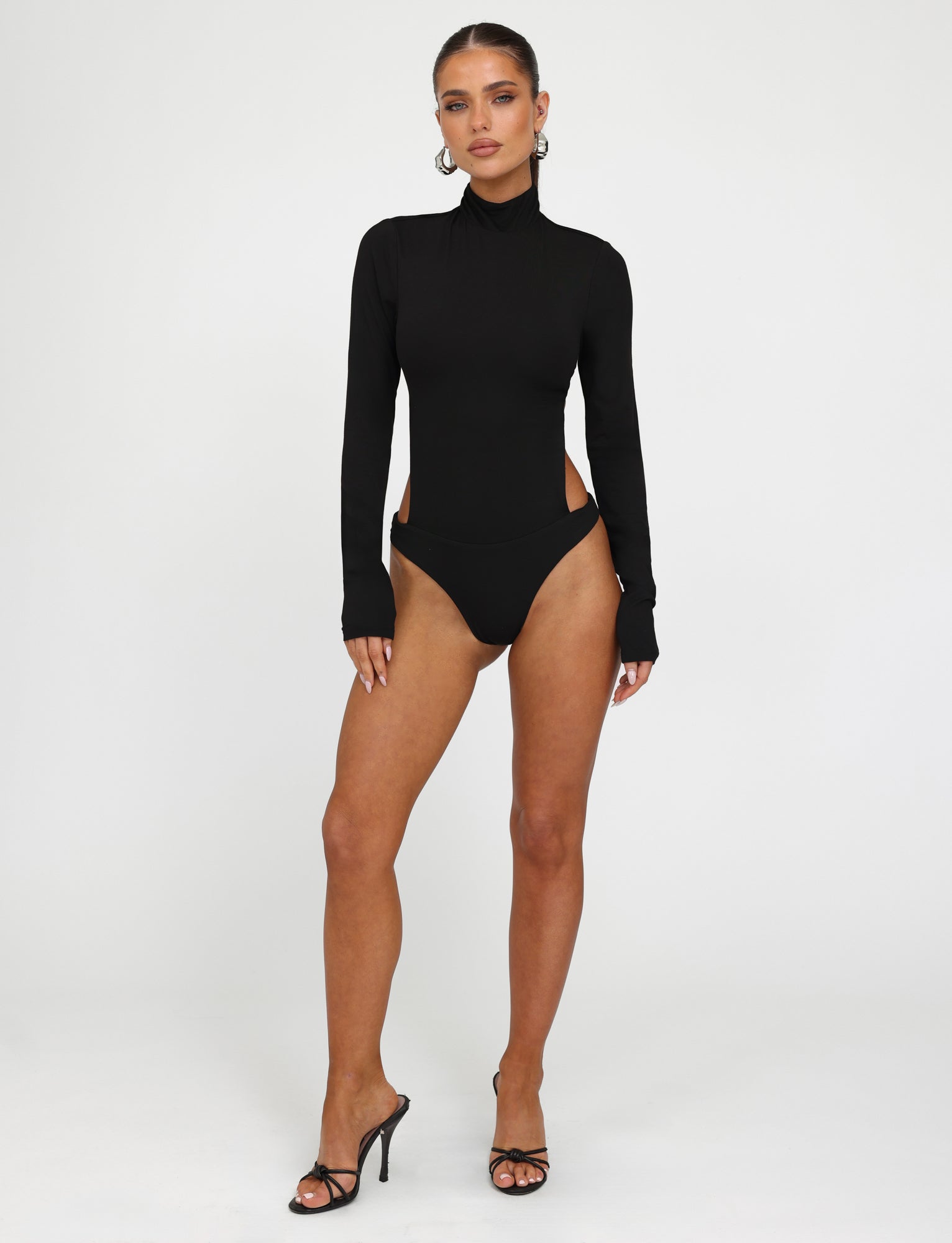 A woman stands confidently against a white background, wearing the AKARI BODYSUIT - BLACK from TIGERMIST, black high-heeled sandals, and large hoop earrings. She has her hair pulled back and poses with her weight shifted to one leg, hands relaxed at her sides.