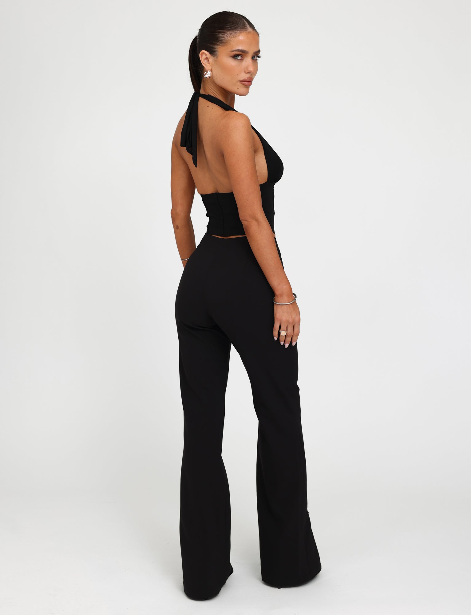 A woman stands facing slightly away from the camera in the chic TIGERMIST DAMIRA Pant, a black halter-top jumpsuit with flared pants. Her hair is tied back in a sleek ponytail, and she accessorizes with bracelets. The background is plain white.