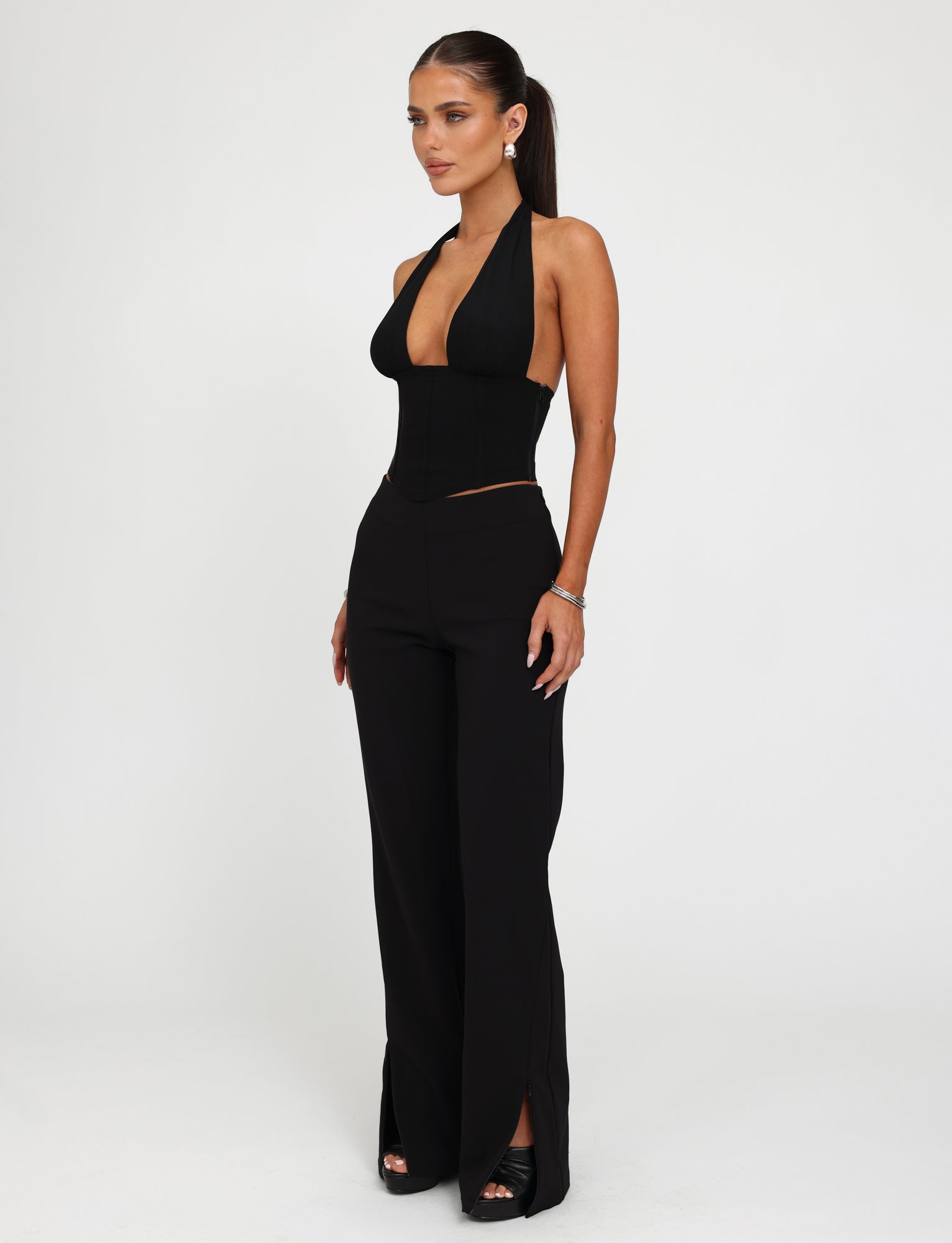 A woman stands against a plain white background, wearing a stylish TIGERMIST outfit consisting of a black halter top with a plunging neckline and the DAMIRA PANT - BLACK, which features high-waisted flared bottoms. Her long hair is tied back in a ponytail, and she completes the look with high-heeled shoes.