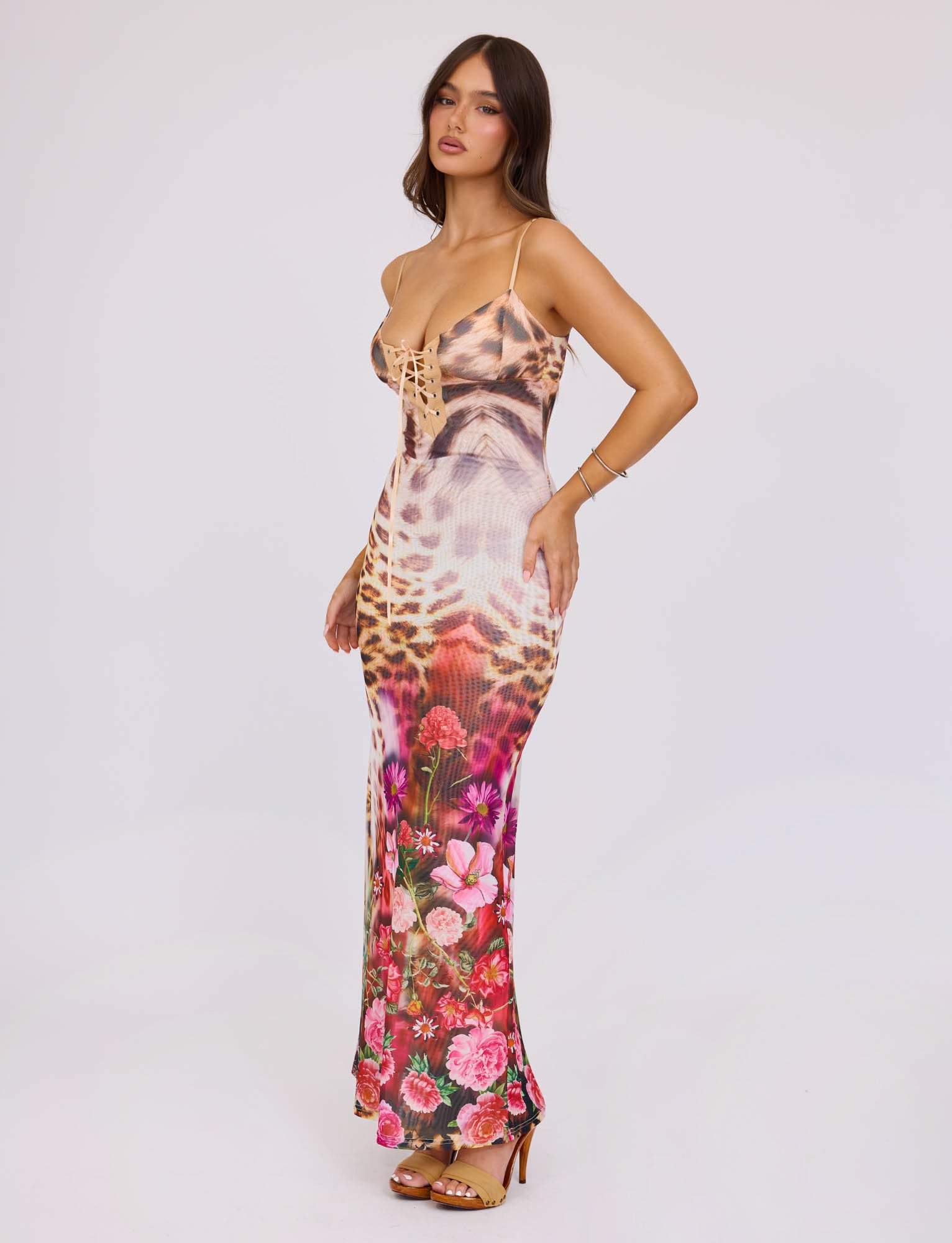 A woman stands against a plain white background, exuding confidence in the TIGERMIST Kalilah Dress - Multi: Leopard, which features a leopard print design on the top half and a vibrant floral pattern on the lower half. With long, dark hair cascading over her shoulders and high-heeled sandals, she embodies modern elegance.