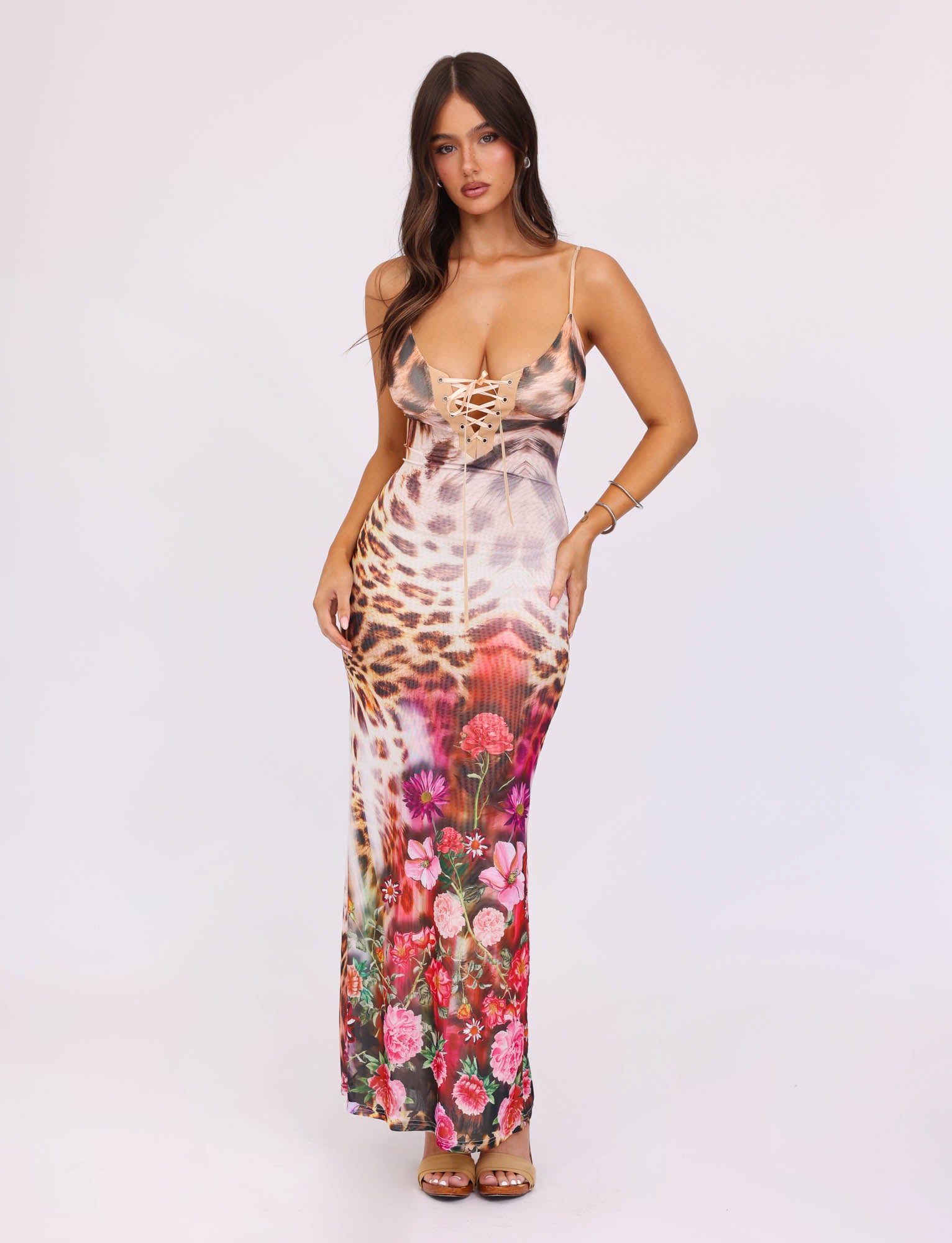 A woman with long dark hair stands against a plain background, wearing a colorful KALILAH DRESS - MULTI : LEOPARD by TIGERMIST. The maxi dress features a mix of animal and floral prints with lace-up details at the bust. She is posing with her hands lightly grasping the sides of her dress.