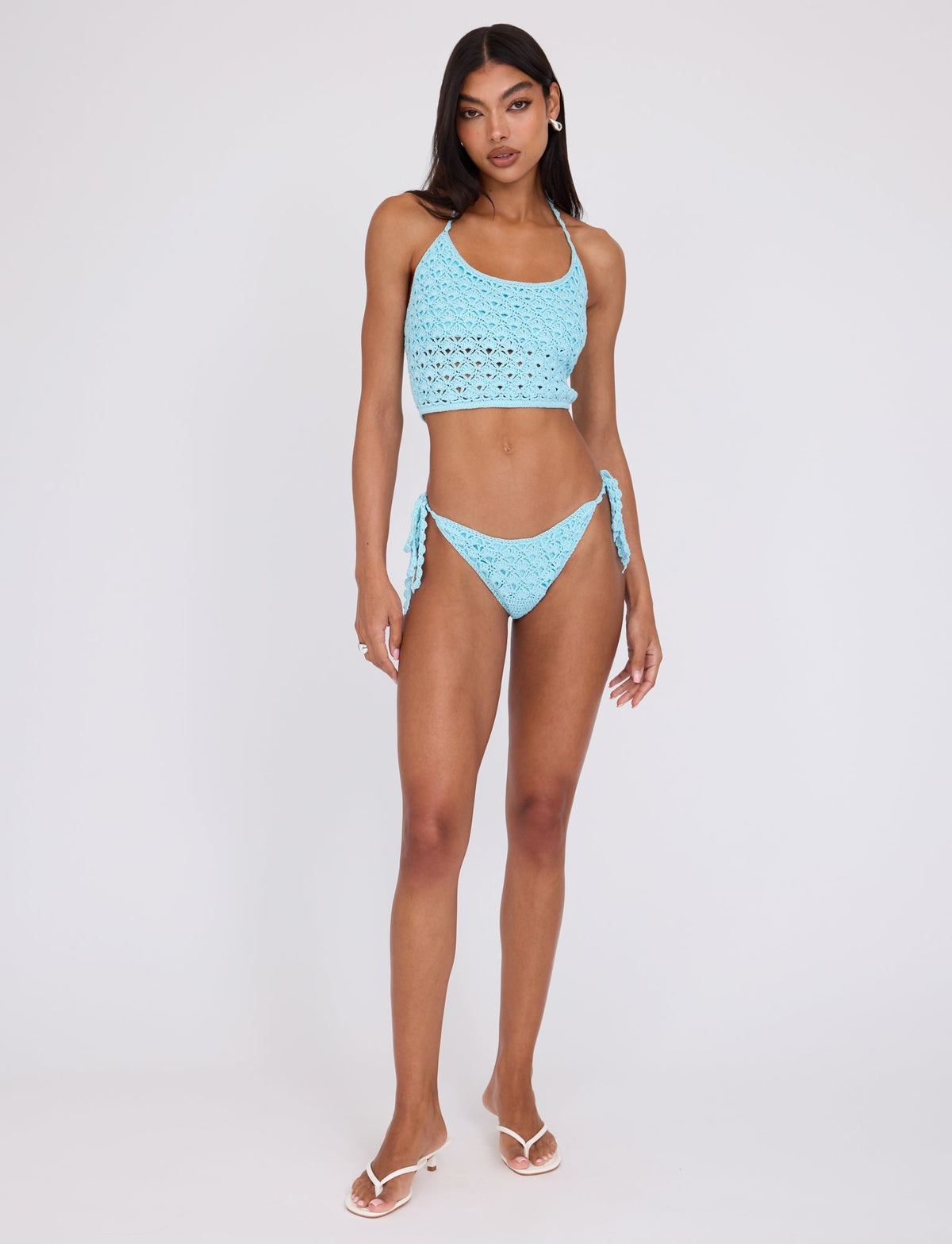 A woman stands against a plain background wearing the TIGERMIST SUNNIVA BIKINI TOP in baby blue. The tank-style bikini top is paired with matching bottoms featuring side ties. She complements her look with white flip-flops, and her hair is styled straight, cascading over her shoulders. She has a neutral expression.