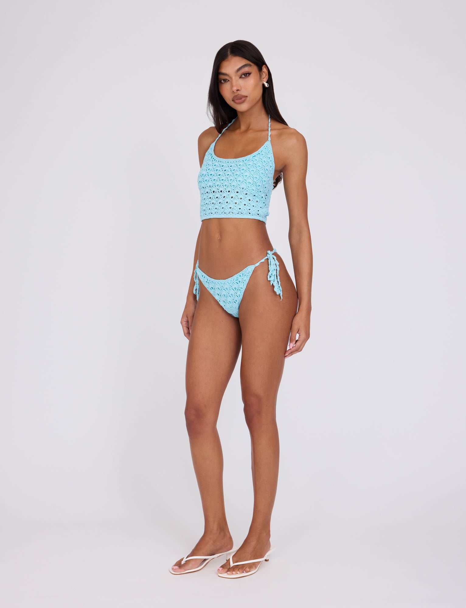 A woman stands against a plain white background, wearing the TIGERMIST SUNNIVA BIKINI TOP in baby blue with a lacy eyelet pattern. She is also wearing white flip-flops and has long, dark hair. She gazes directly at the camera with her hands resting loosely by her sides.