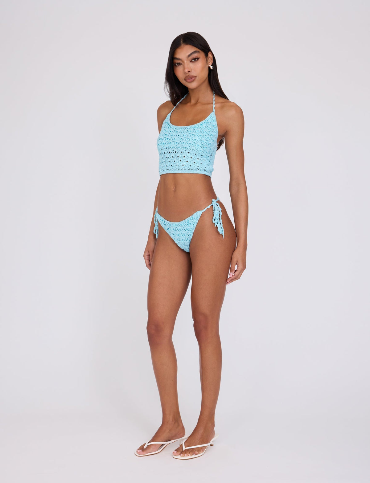 A woman stands against a plain white background, wearing the TIGERMIST SUNNIVA BIKINI TOP in baby blue with a lacy eyelet pattern. She is also wearing white flip-flops and has long, dark hair. She gazes directly at the camera with her hands resting loosely by her sides.