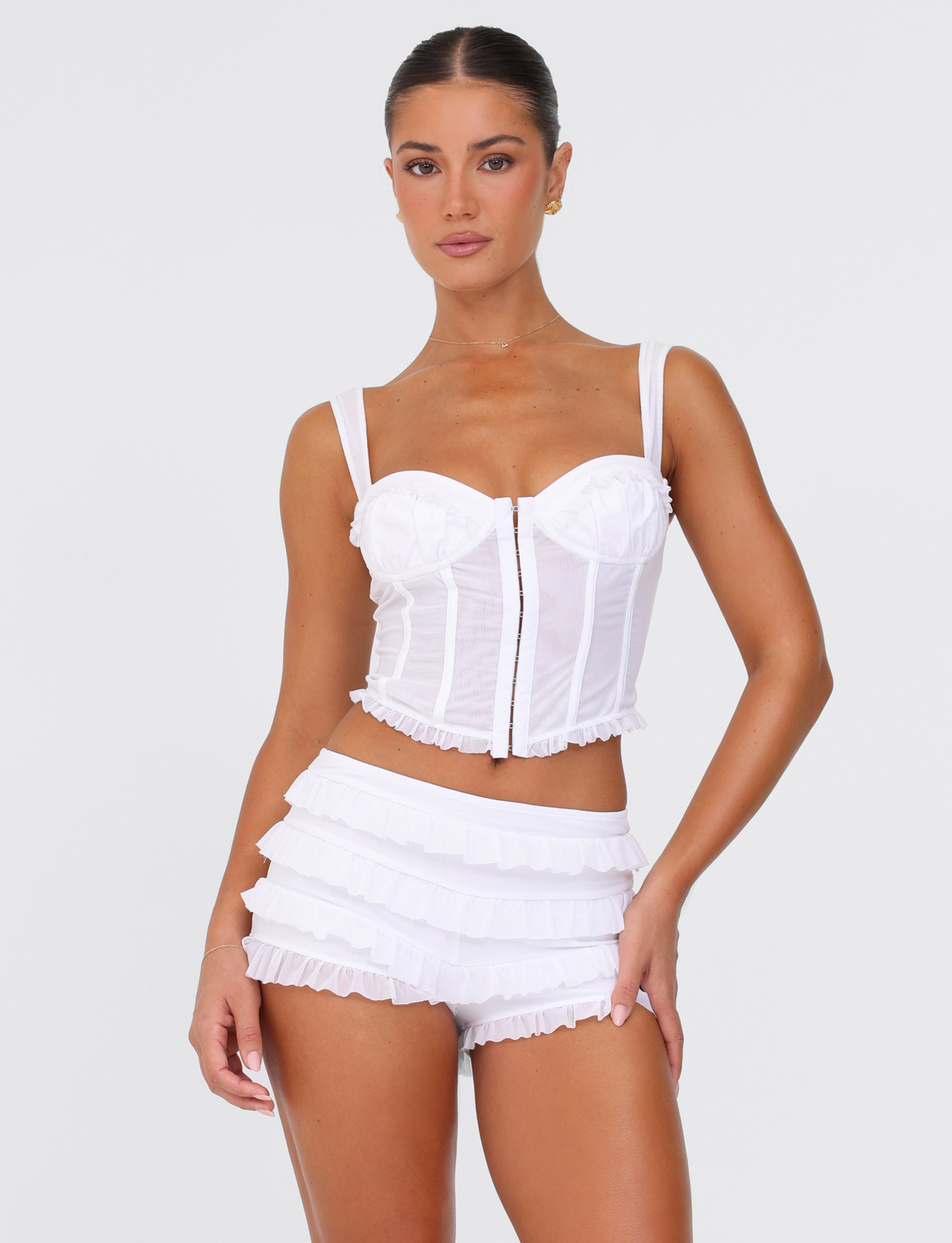 A woman with her hair tied back poses confidently in a TIGERMIST GINNY TOP - WHITE : OPTIC WHITE, a fitted corset-style top with a front zipper, paired with matching high-waisted ruffled shorts against a plain white background.