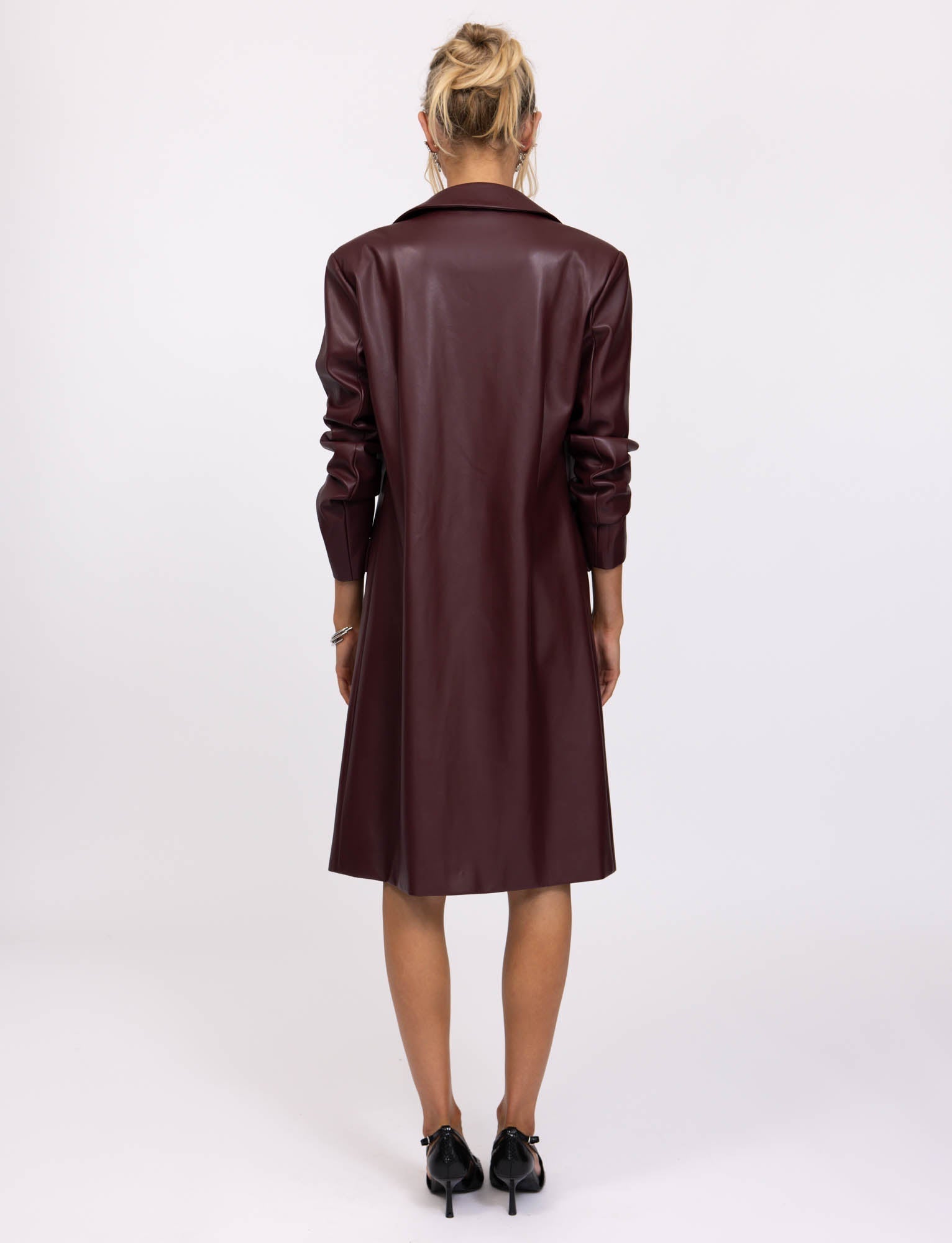 A person with blonde hair is standing facing away, wearing the Cherry Red Lyon Trench Coat from TigerMist. The coat has a smooth finish and reaches just above the knees. The person is also wearing black heeled shoes. The background is plain and white.