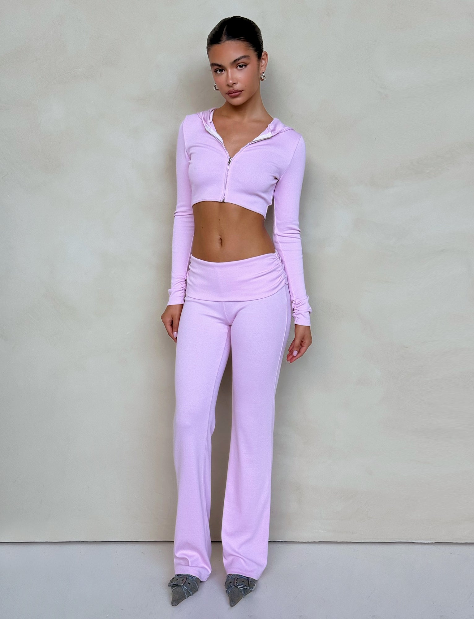 A person stands against a neutral wall wearing a light pink, long-sleeved, cropped zip-up hoodie and the KARINA PANT in Blush from TIGERMIST. They are looking directly at the camera with a neutral expression. The outfit is complemented by pointed-toe shoes.