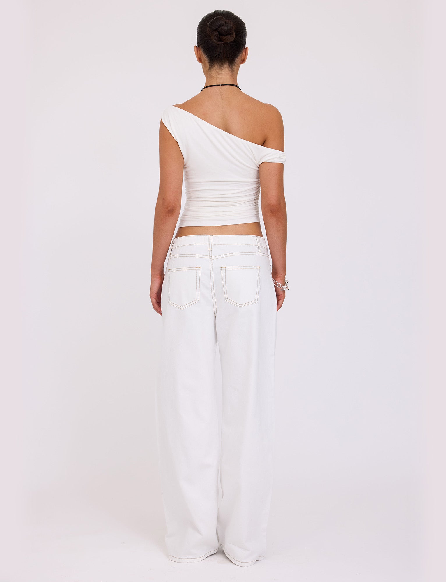 A person with hair in a bun is standing with their back to the camera, wearing a one-shoulder white top and loose FELIX JEAN - WHITE pants from TIGERMIST with visible pockets. The background is plain white.