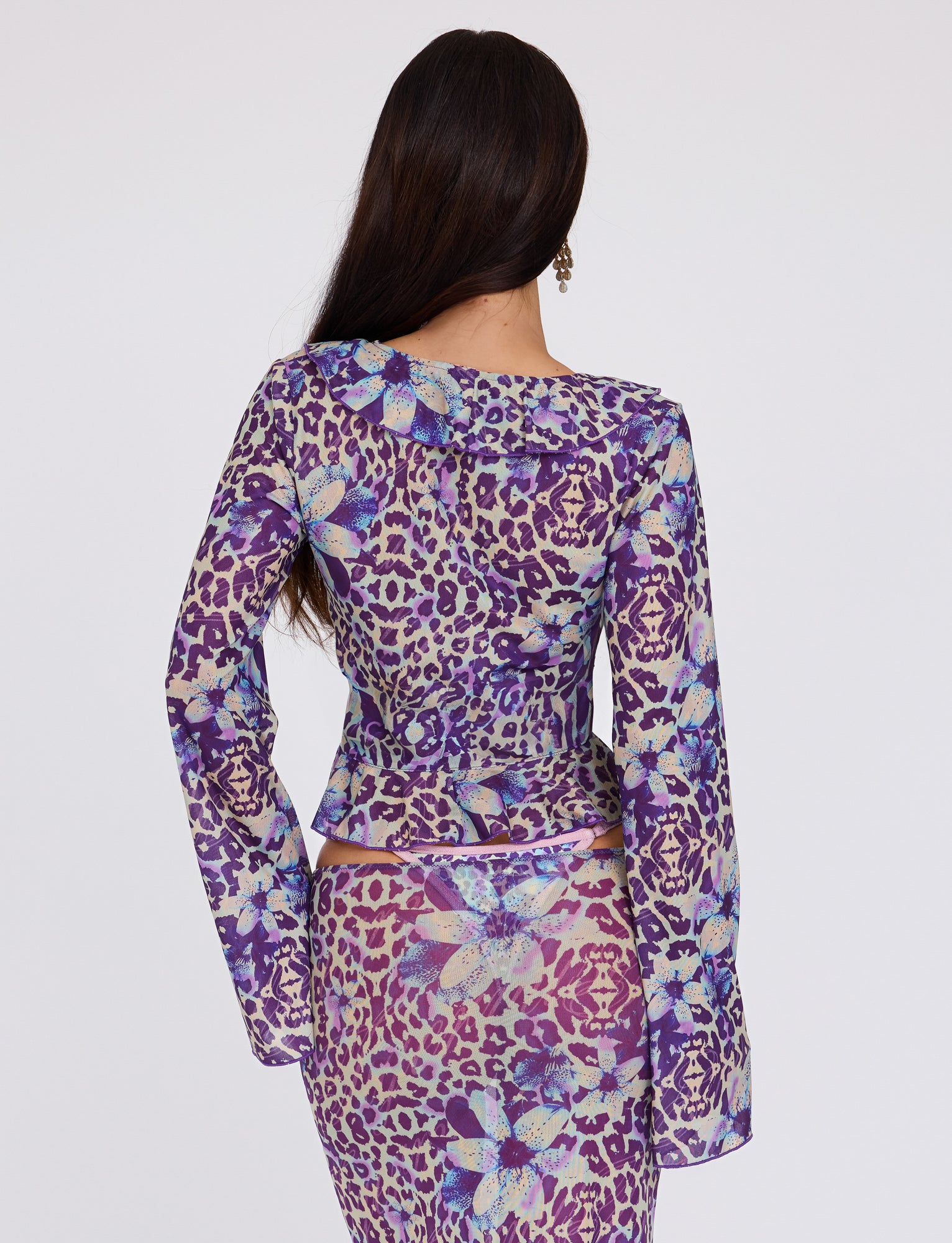 A woman with long dark hair faces away from the camera, modeling the CORVIN MESH TOP - BLUE : BY POPPY LEOPARD from TIGERMIST. The form-fitting top, featuring a vibrant purple leopard print and large blue flowers, stands out brilliantly against the white background.