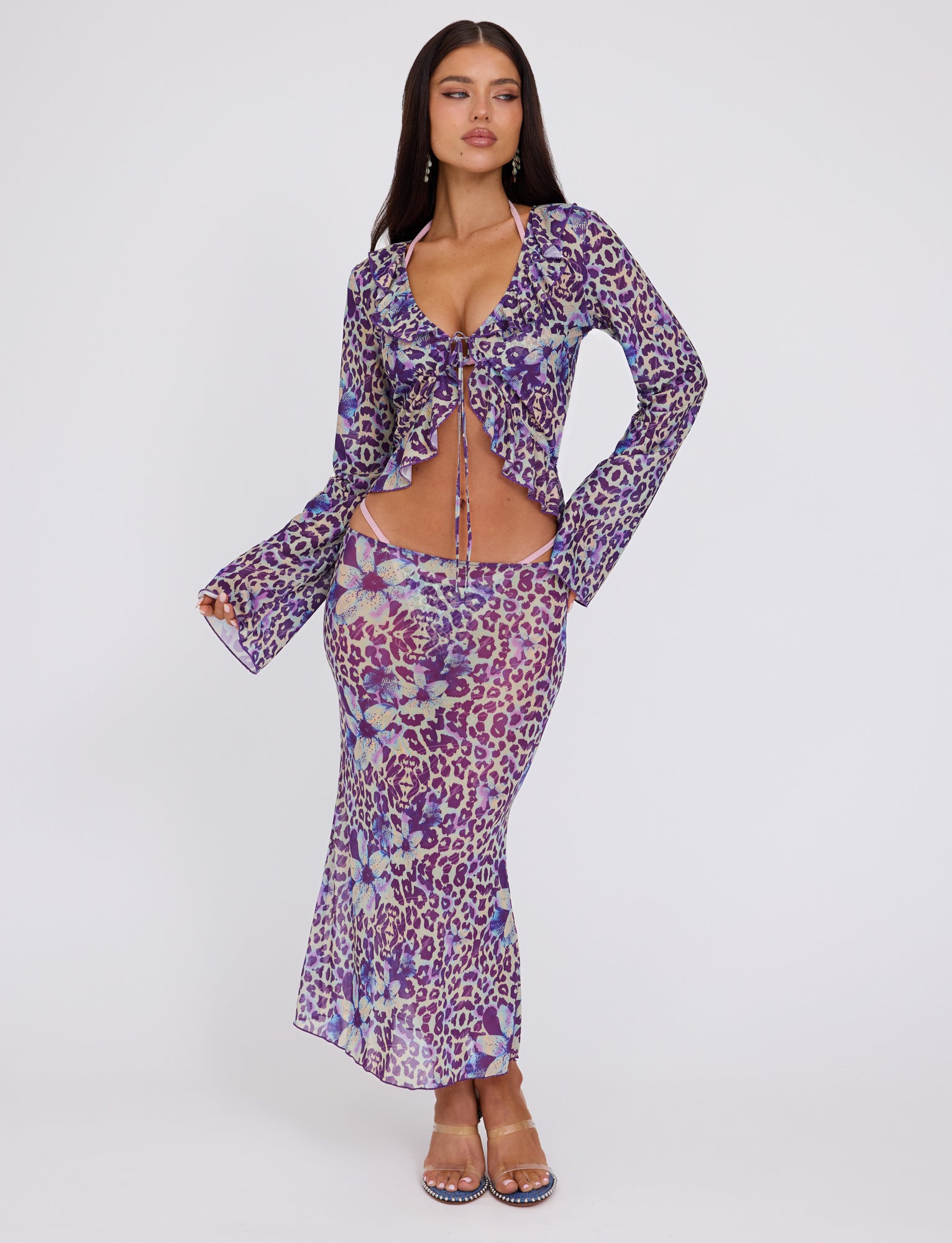 A woman with long hair is standing against a plain background, wearing the CORVIN MESH TOP - BLUE : BY POPPY LEOPARD from TIGERMIST. The form-fitting, long-sleeve top has a deep V-neck and string ties at the front, showcasing a vibrant purple and blue leopard print. She completes her look with high-heeled sandals.