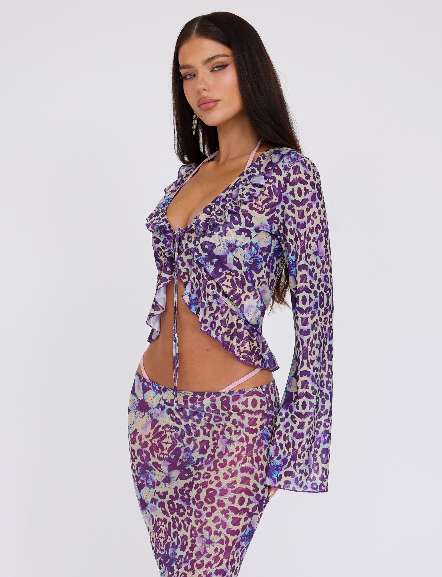 A woman with long, straight dark hair confidently poses against a plain white background, donning a TIGERMIST stylish outfit. She wears the CORVIN MESH TOP - BLUE, which features a chic purple and lavender leopard print with a tie front, paired with the matching skirt.