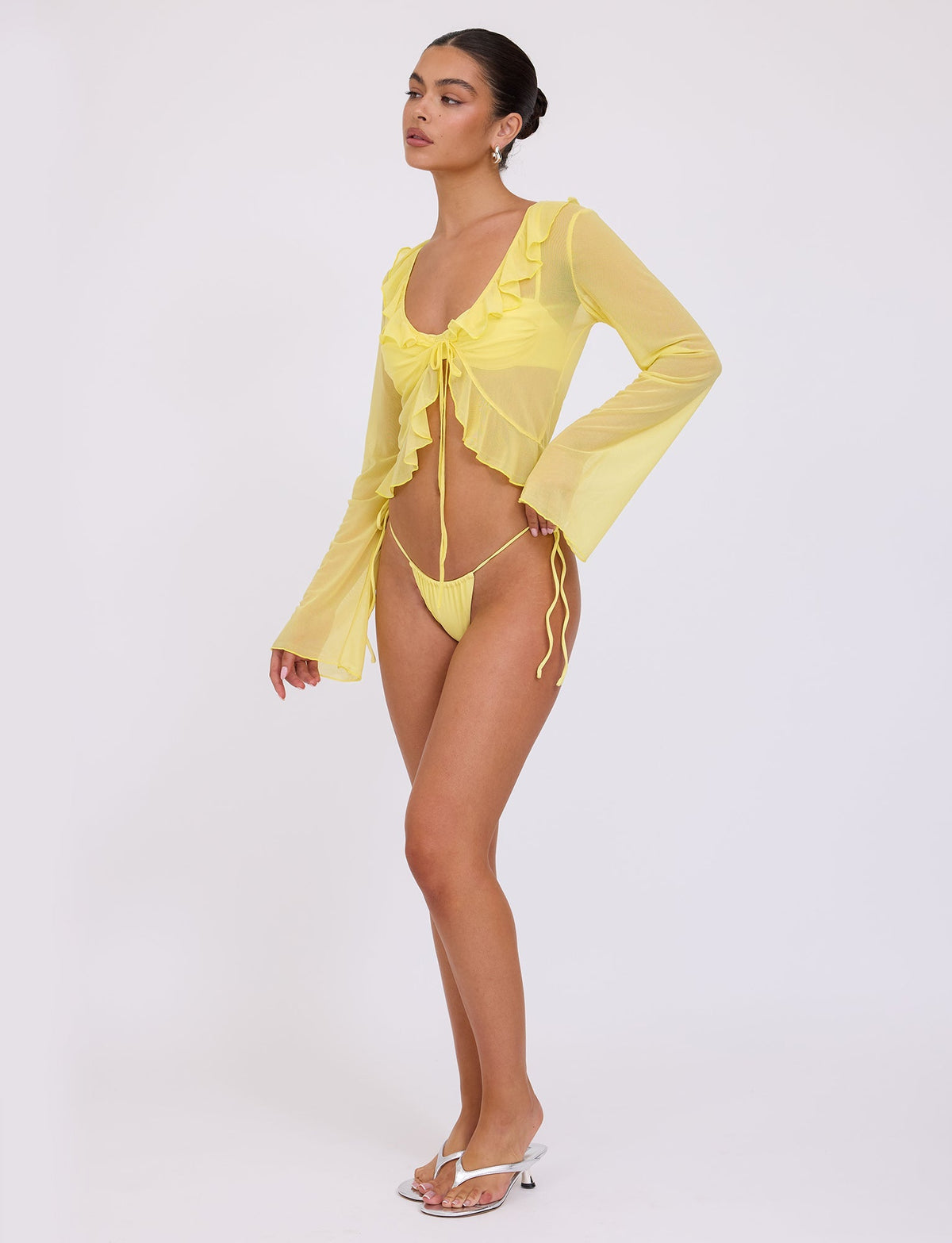A woman stands in front of a plain white background wearing a sheer, long-sleeved TIGERMIST CORVIN TOP in Yellow Lemon with a ruffle neckline and matching yellow tie-side bikini bottoms. She is also wearing white flip-flops and small hoop earrings, with her hair pulled back in a sleek bun.