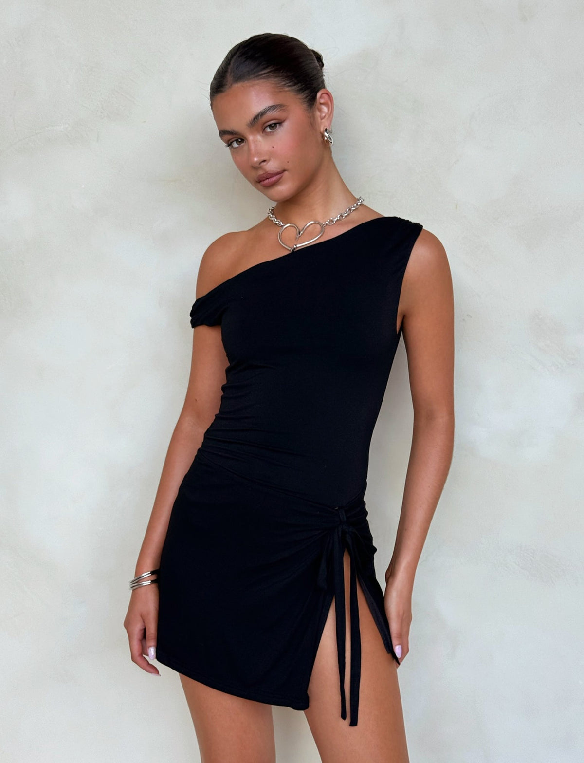 A person with dark hair stands against a light background, wearing the KADENCE DRESS - BLACK by TIGERMIST. The black one-shoulder dress has a knot detail on the side and an asymmetrical neckline, featuring a high split that adds to its elegance. They accessorize with silver hoop earrings, a heart-shaped necklace, and a bracelet on their left wrist.