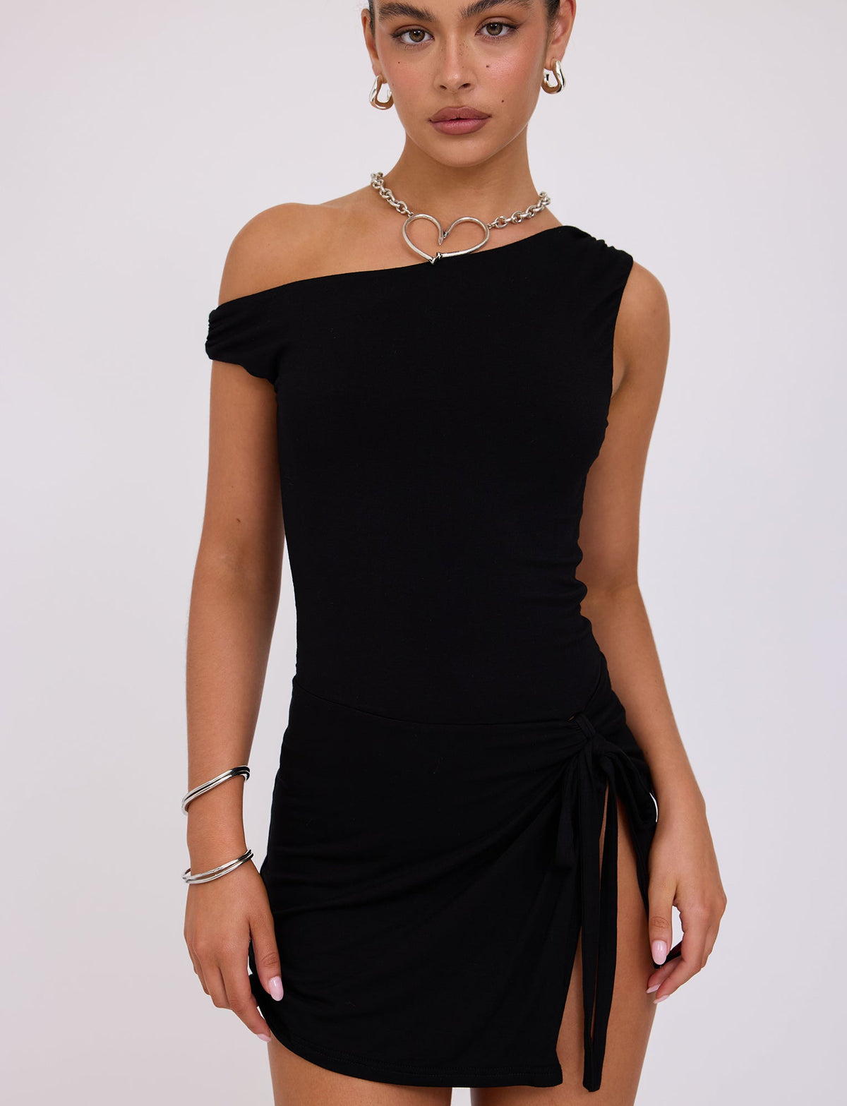 A person wears the KADENCE DRESS - BLACK by TIGERMIST, which features an asymmetrical neckline, a side tie detail, and a thigh-high slit. The look is complemented by a heart-shaped necklace, hoop earrings, and silver bracelets. Standing against a plain background, the person gazes directly at the camera.