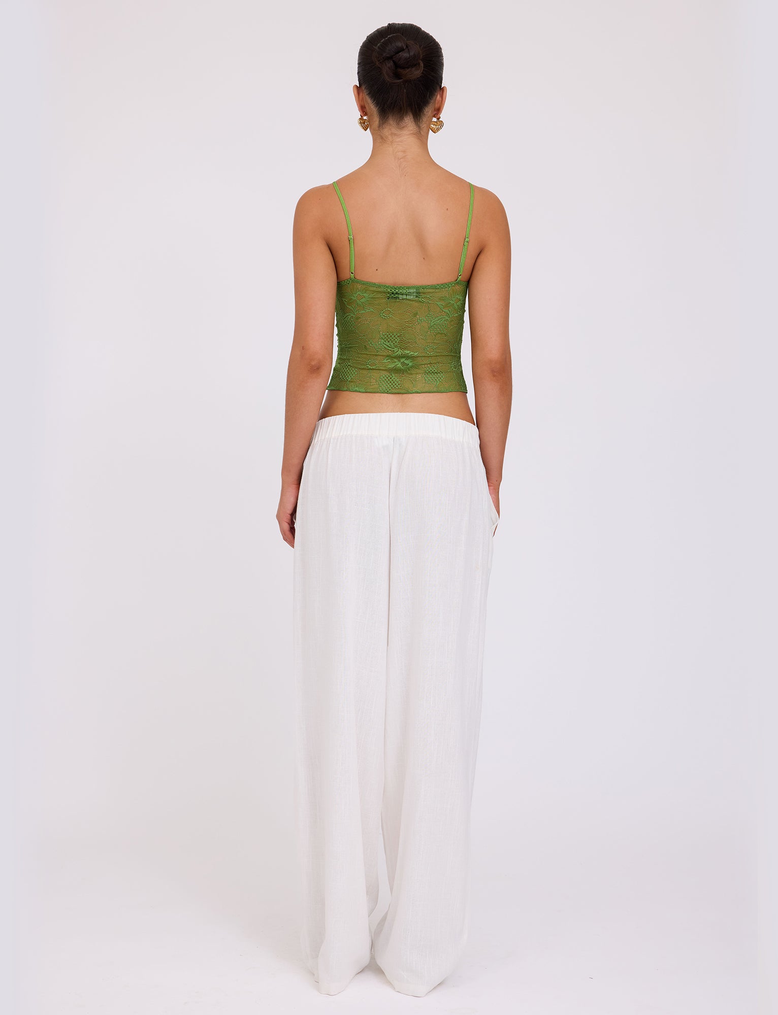 A person with their hair in a bun is standing and facing away from the camera, showcasing an ensemble that could be straight out of a TIGERMIST collection: the SASKIA TOP in green paired with loose, white pants. The background is white and plain.