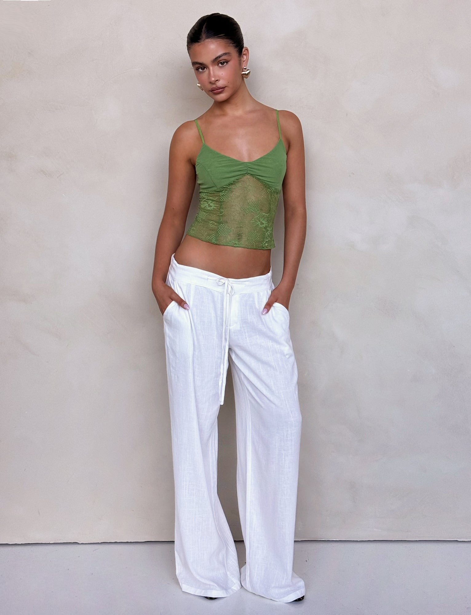 A person wearing the SASKIA TOP in a light green, semi-transparent fabric from TIGERMIST, paired with white wide-legged pants, stands against a plain, light-colored background. Their hands are in their pockets, and they have a neutral facial expression.