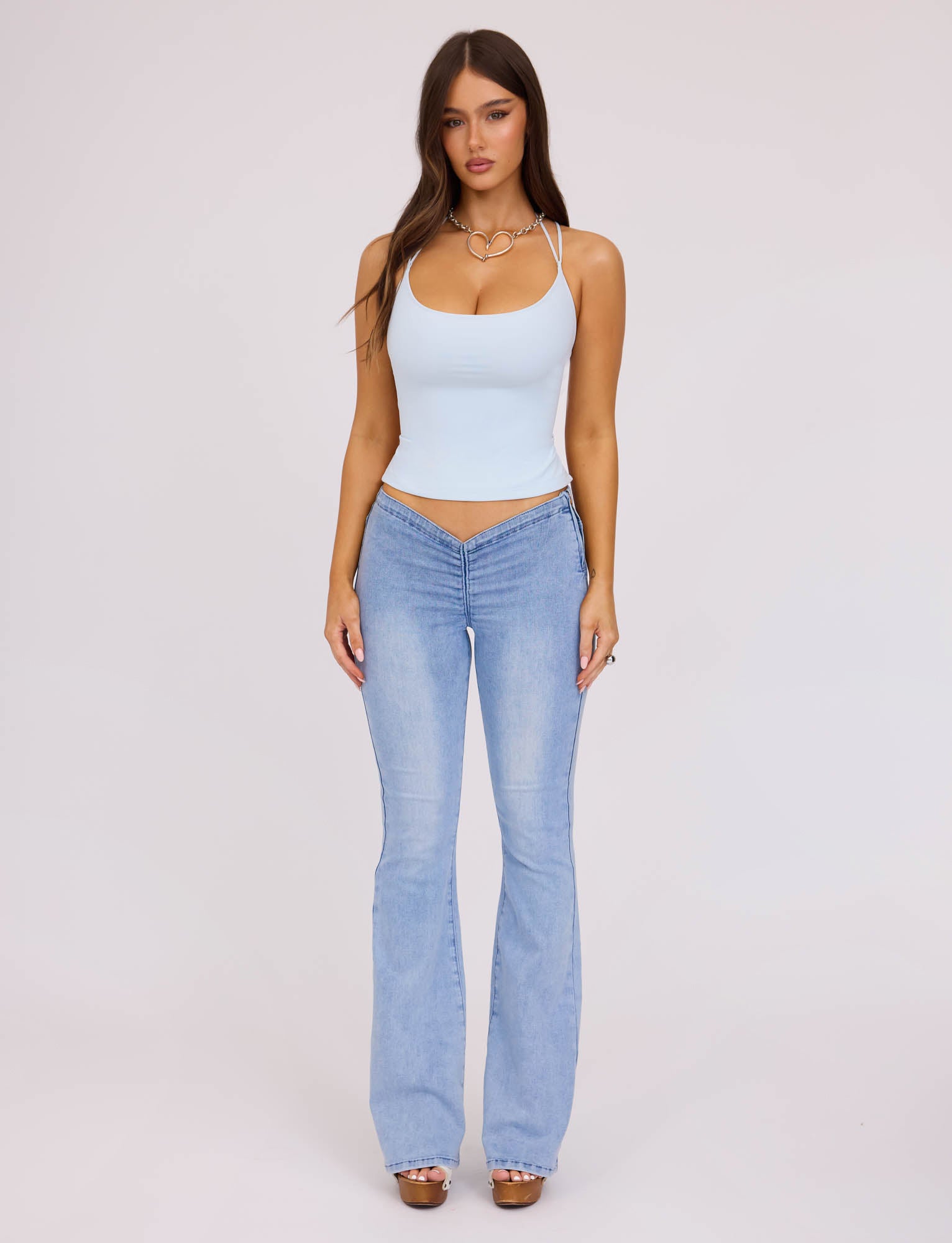 A woman with long hair stands against a plain white background, wearing the ZAIDA TOP - PASTEL BLUE and matching light blue flared jeans from TIGERMIST. She accessorizes with a necklace and open-toe shoes. Her hands are relaxed by her sides, and she looks directly at the camera.