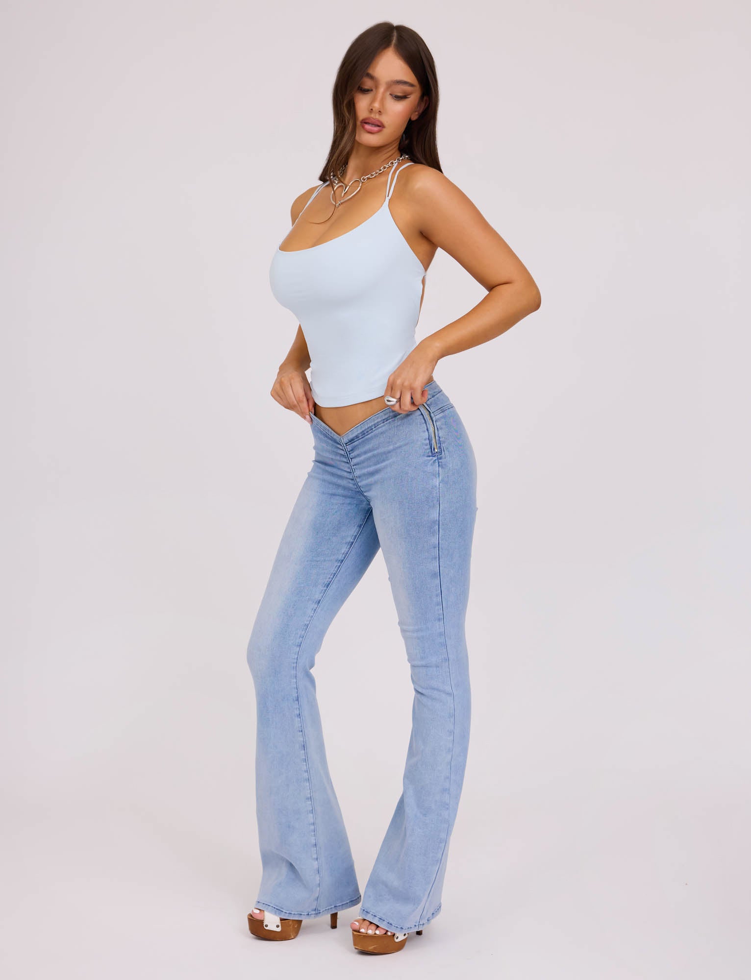 A woman with long dark hair stands against a plain background, dressed in a light blue, form-fitting ZAIDA TOP - BLUE from TIGERMIST and light-wash, flared jeans. She poses with her hands resting on the waistband of her jeans and wears white high-heeled shoes.