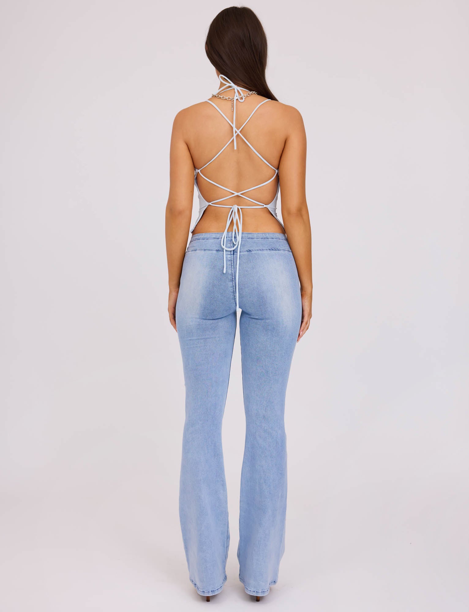A person with long, dark hair is standing with their back to the camera, showcasing their stylish Tiger Mist ensemble. They are wearing light blue, high-waisted jeans that flare at the bottom and a white, strappy, backless ZAIDA TOP in pastel blue from TIGERMIST. The background is plain white.