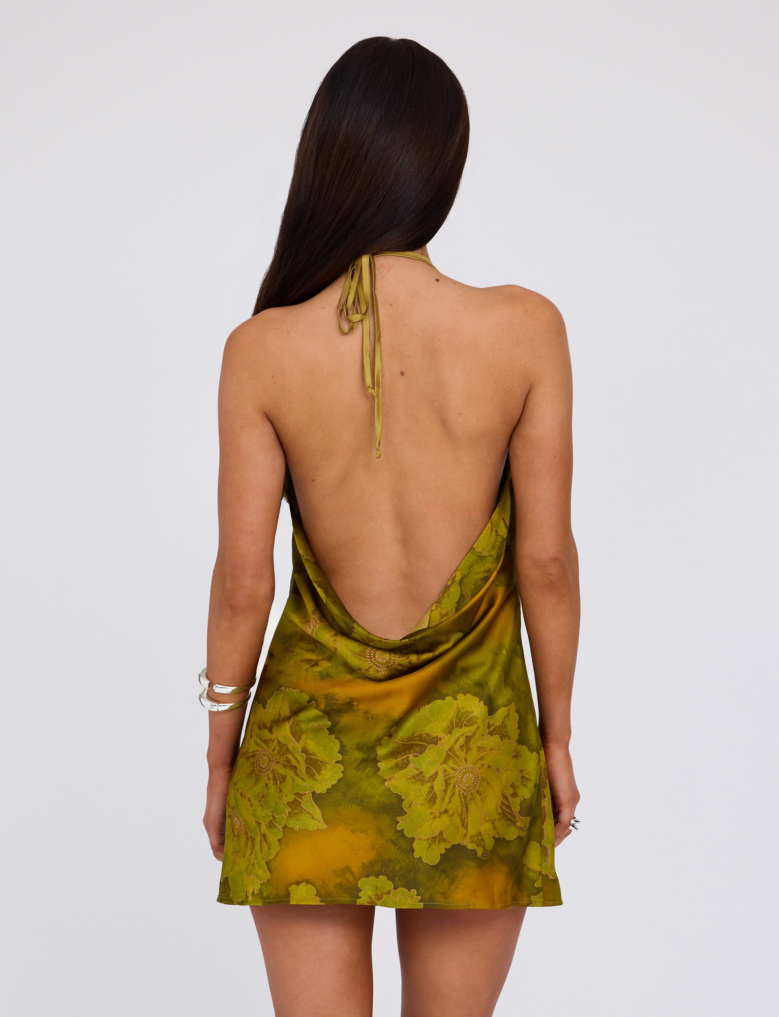 A woman with long brown hair, seen from behind, wears the TIGERMIST MONROE DRESS - GREEN in Water Colour Floral, a halter mini dress with a low open back. She accessorizes with bracelets and poses against a plain light background.