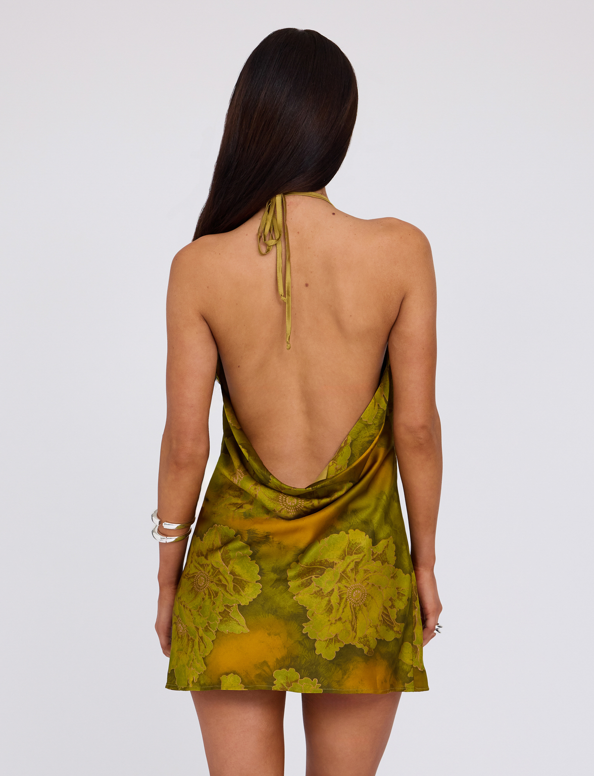 A woman with long brown hair, seen from behind, wears the TIGERMIST MONROE DRESS - GREEN in Water Colour Floral, a halter mini dress with a low open back. She accessorizes with bracelets and poses against a plain light background.