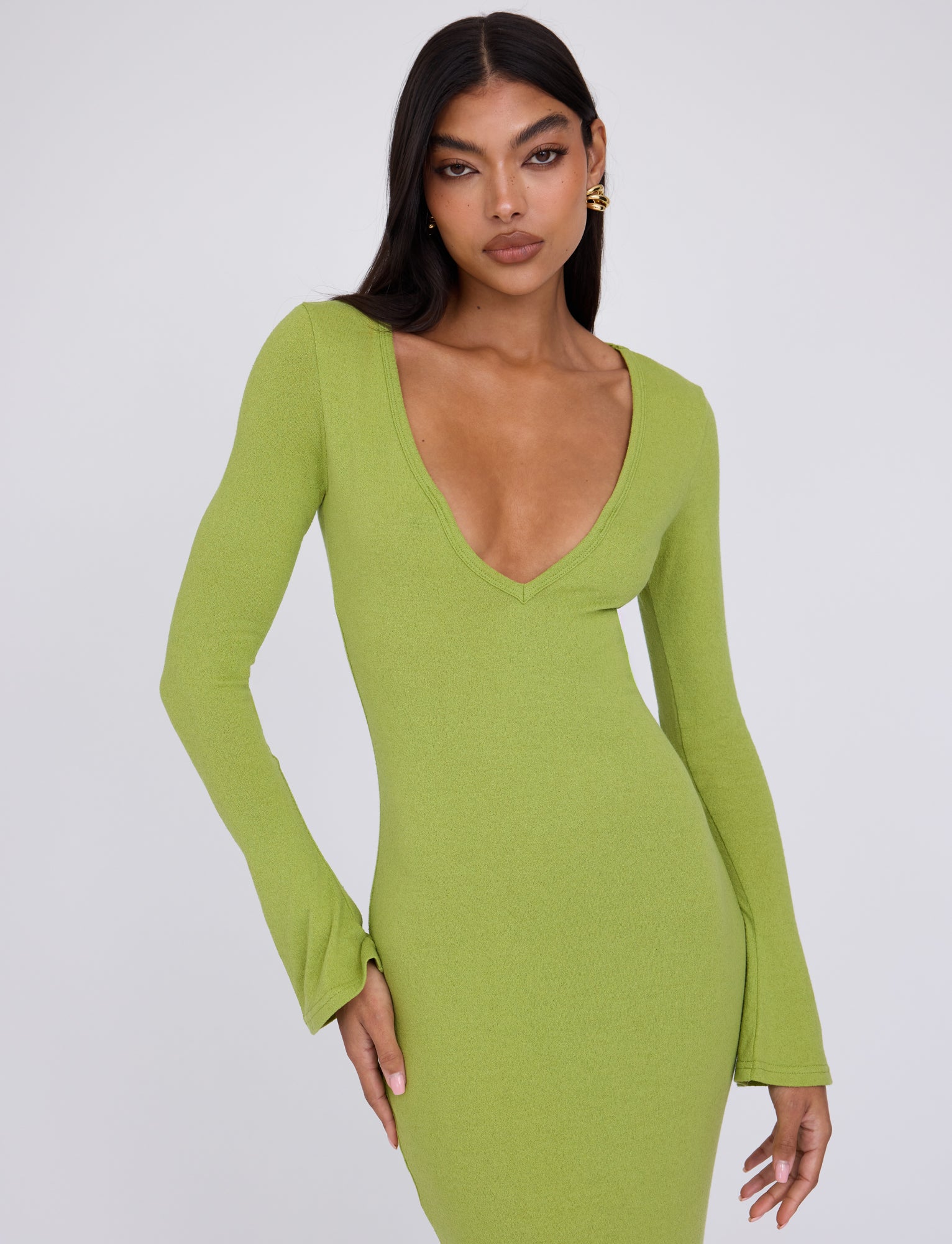 A person with long dark hair is wearing the ROMIE DRESS in Matcha Green by TIGERMIST, which features a fitted design, long sleeves, and a deep V-neckline. The individual poses with one hand on their hip and the other arm down by their side, against a plain white backdrop.
