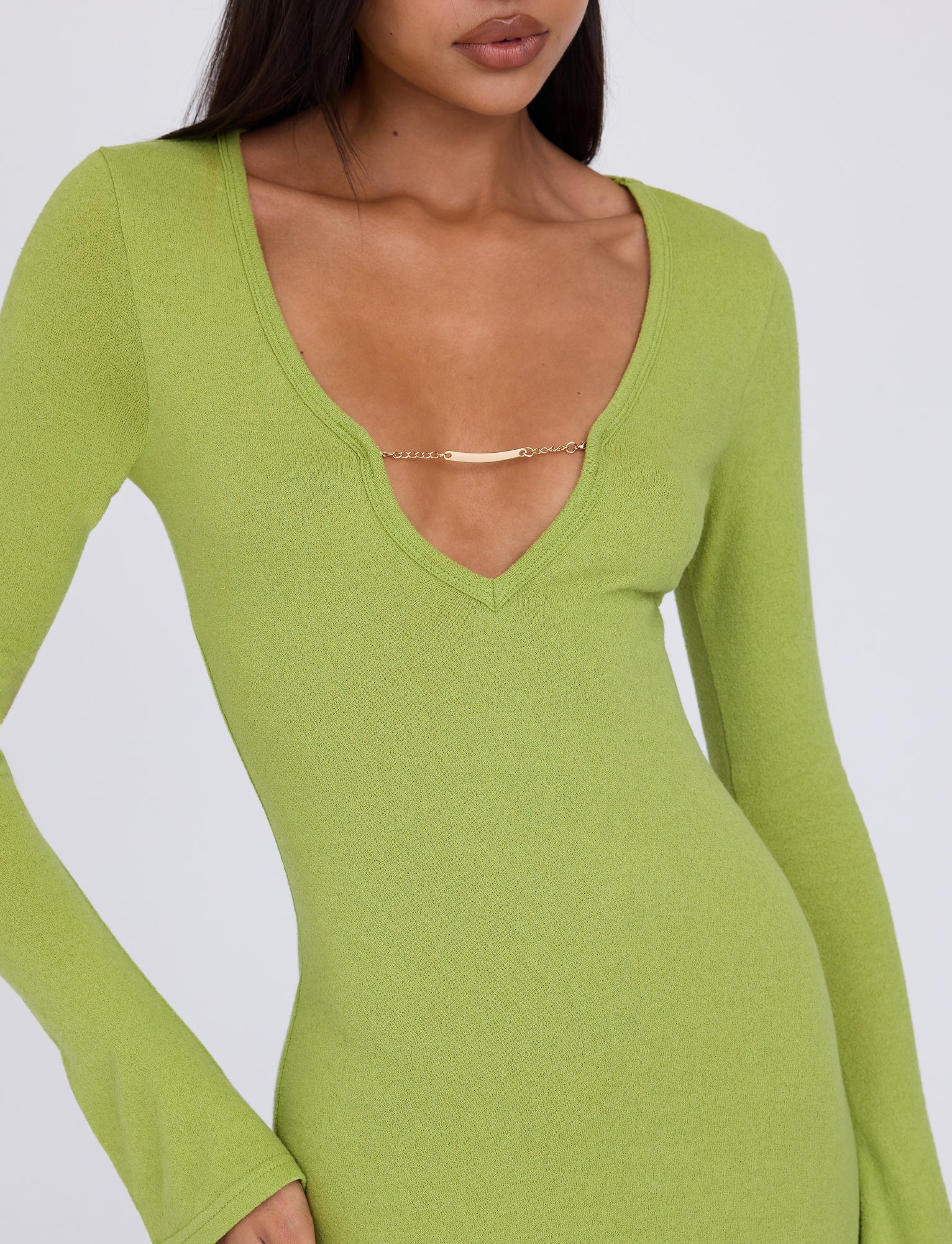 A person is wearing the Romie Dress in Matcha Green from TigerMist. The long-sleeve dress with a deep V-neck boasts a decorative gold pin at the neckline's sides. The individual's hands are relaxed by their sides, against a plain white background.