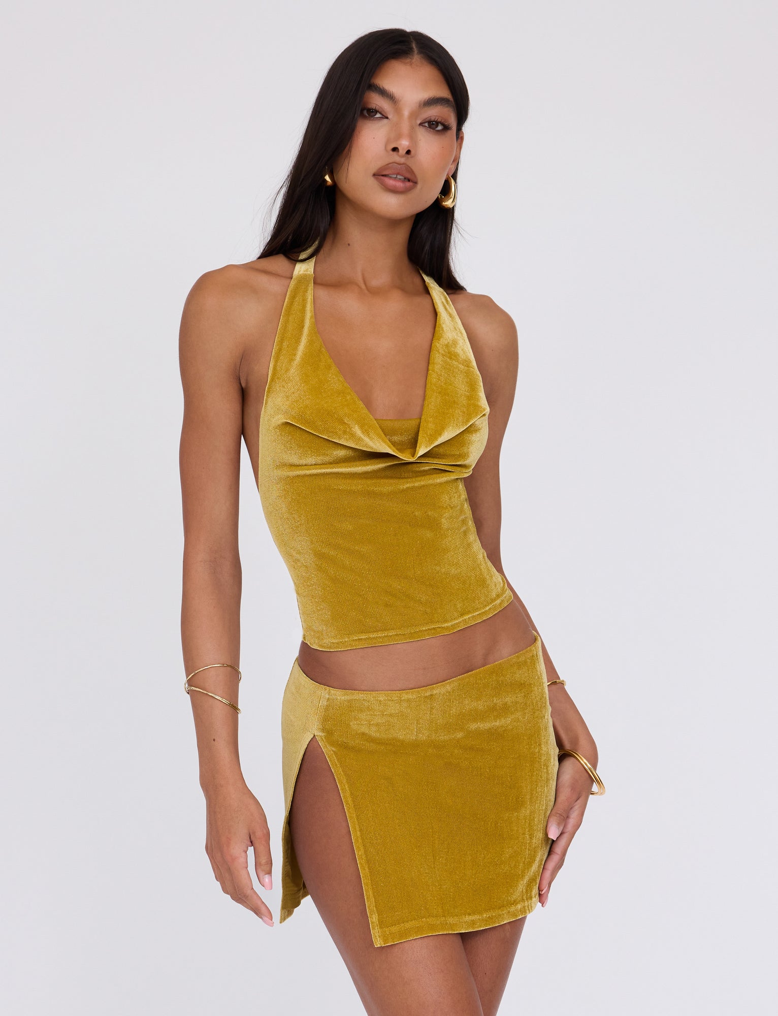 A woman with long dark hair models the HARLOWE TOP - GOLD by TIGERMIST against a plain background. The ensemble features a mustard velvet halter top with a draped neckline and a matching mini skirt with a thigh-high slit. She accessorizes with gold hoop earrings and delicate bracelets.