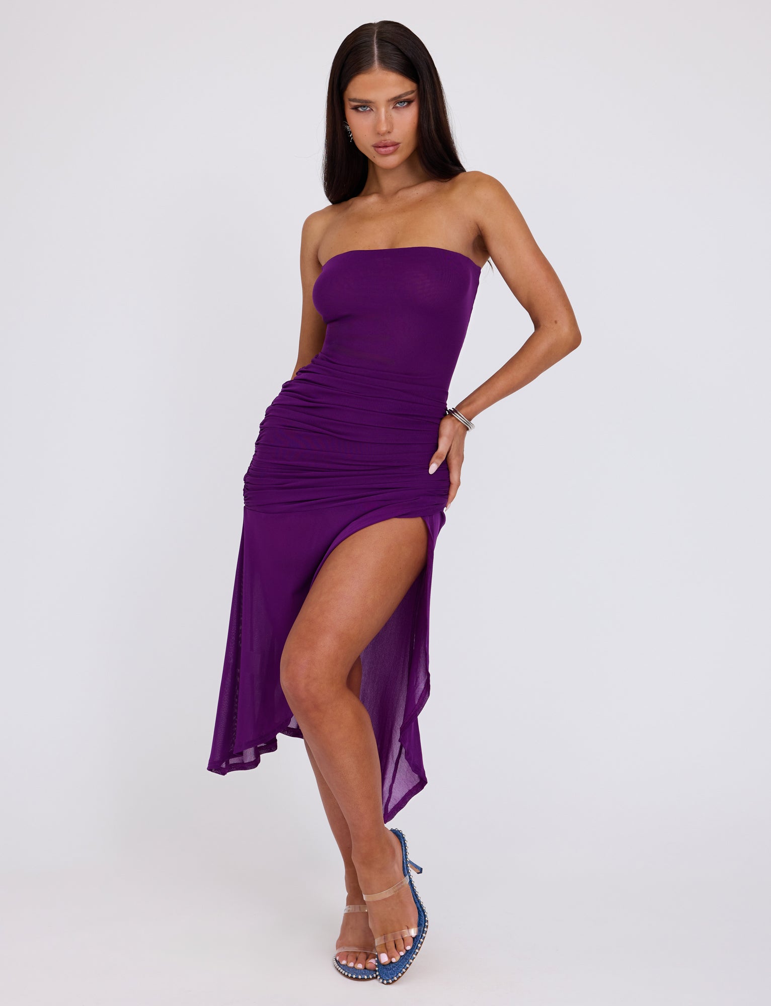 A woman with long, straight hair poses confidently in the TIGERMIST COVE DRESS - PURPLE. The strapless dress features a high slit on one side and a draped design. She wears clear high-heeled sandals with blue accents and has one hand on her hip, striking a pose against a plain background.