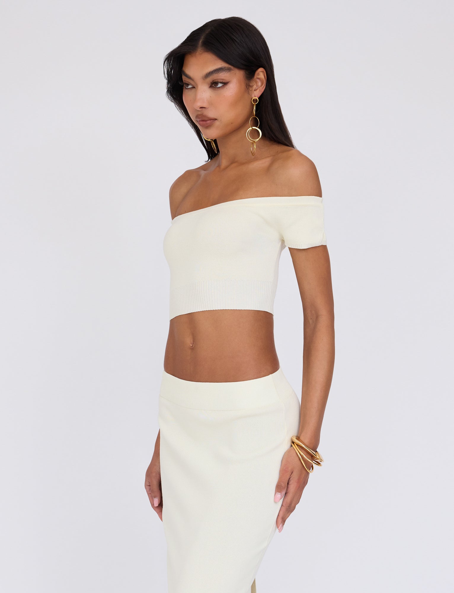 A woman with long dark hair wearing a light-colored, off-the-shoulder crop top and matching skirt from TIGERMIST's LEDGER TOP - WHITE : CREAM collection stands against a plain background. She accessorizes with dangling earrings and multiple bracelets, appearing to look slightly to her left.