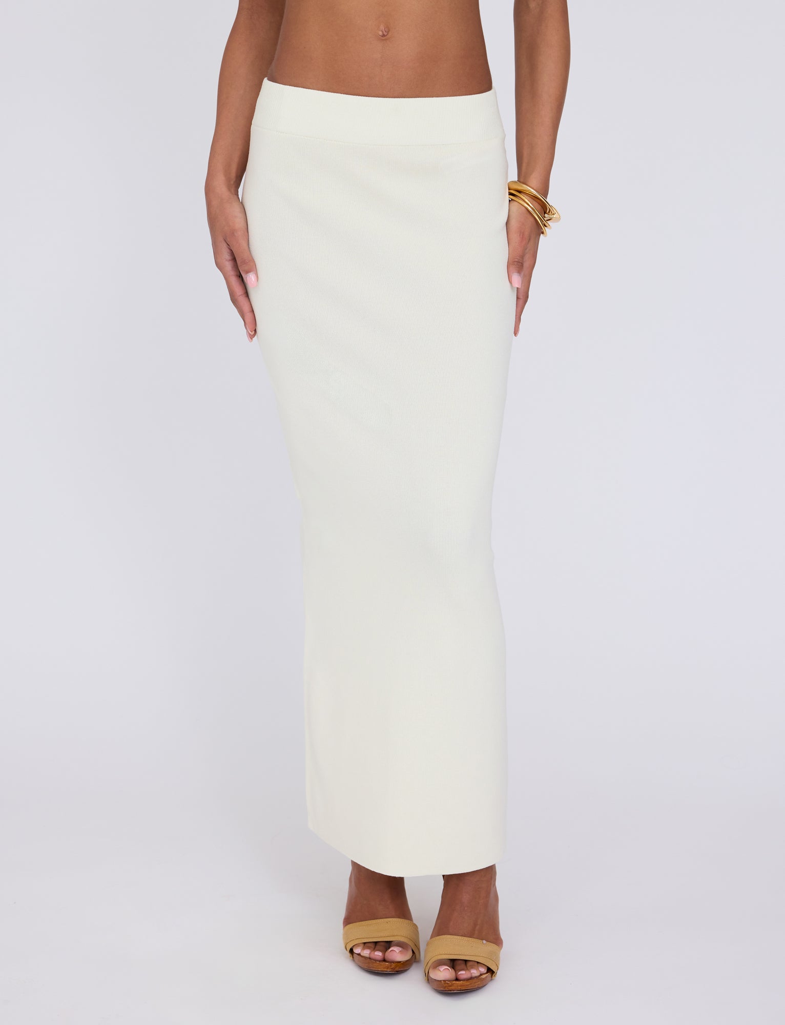 A person wearing the LEDGER SKIRT in white and cream from TIGERMIST, paired with tan open-toed high heels. They are accessorized with gold bangles on their wrist. The photo captures the lower half of the person, from the waist down, against a plain white background.