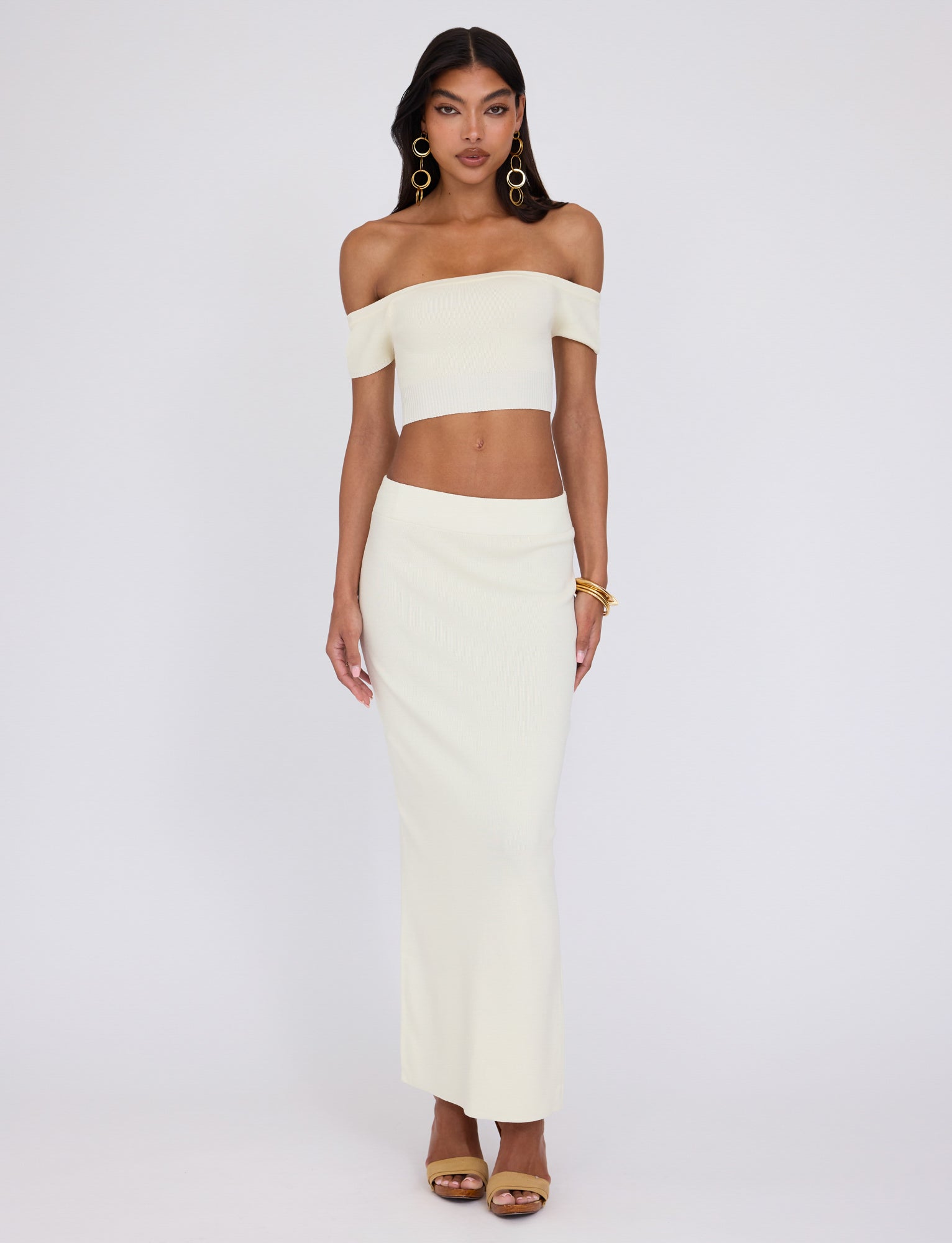 A woman is posing against a plain background, wearing an off-the-shoulder, cream-colored crop top and the high-waisted LEDGER SKIRT - WHITE : CREAM from TIGERMIST. She accessorizes with large hoop earrings, gold bangles, and golden high-heeled sandals and has long, dark hair.
