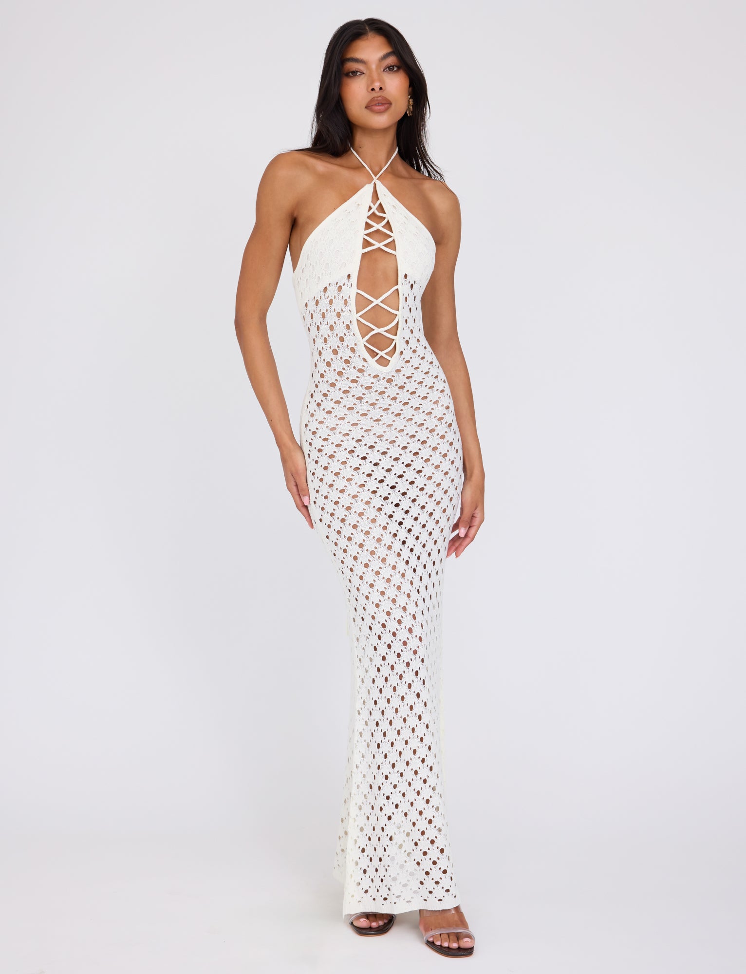 A woman stands against a plain white background, wearing the CIARAN DRESS - WHITE by TIGERMIST. The dress features a long, sleeveless halter-neck design with a lattice pattern and a deep crisscrossed cutout in the front. She has long dark hair and pairs it with brown high-heeled shoes.