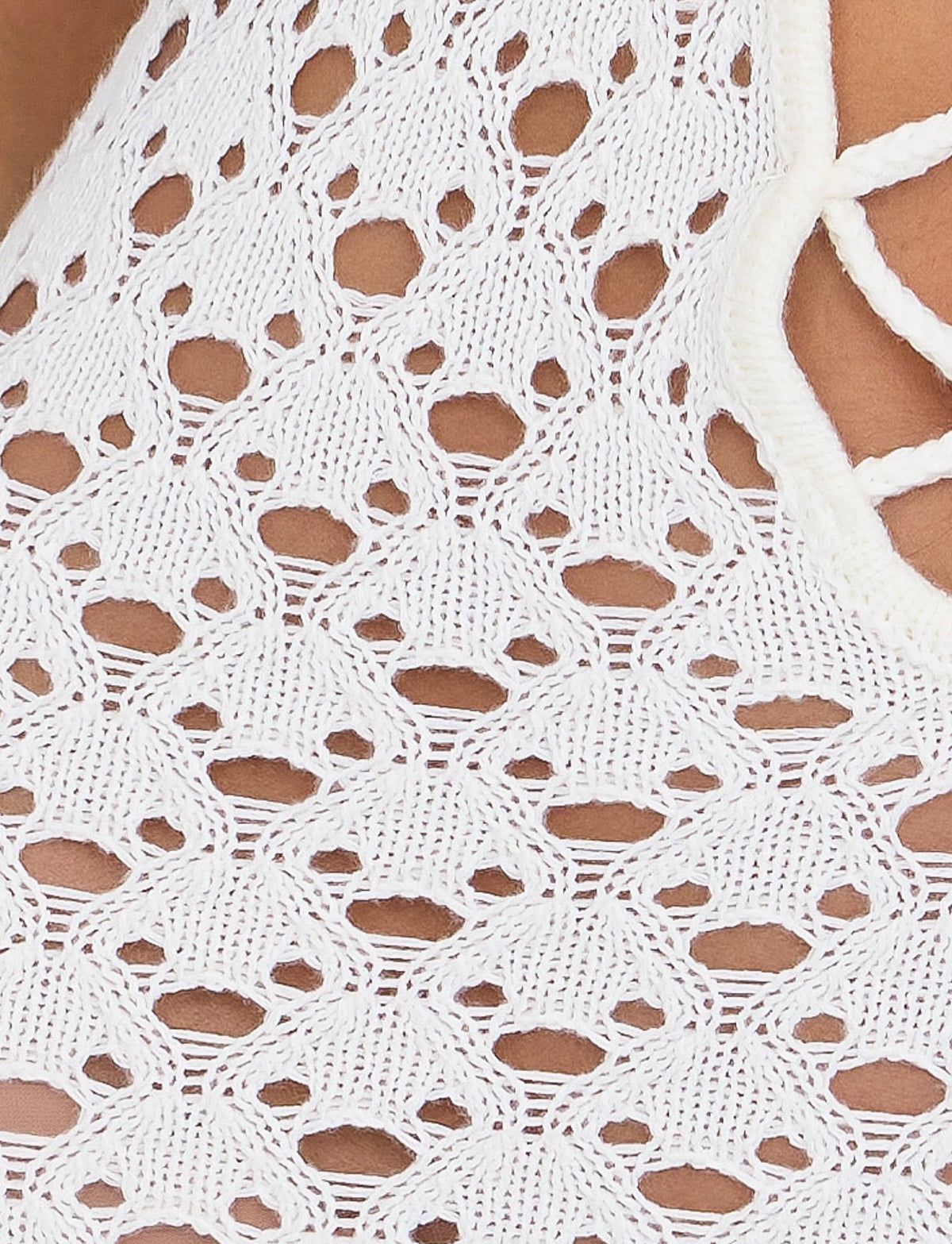 Close-up of a white CIARAN DRESS from TIGERMIST, showcasing its crochet-patterned fabric with an intricate design featuring interlocking semi-circular shapes and small holes. The texture appears delicate and handcrafted. A portion of a TIGERMIST strap is also visible on the right side.